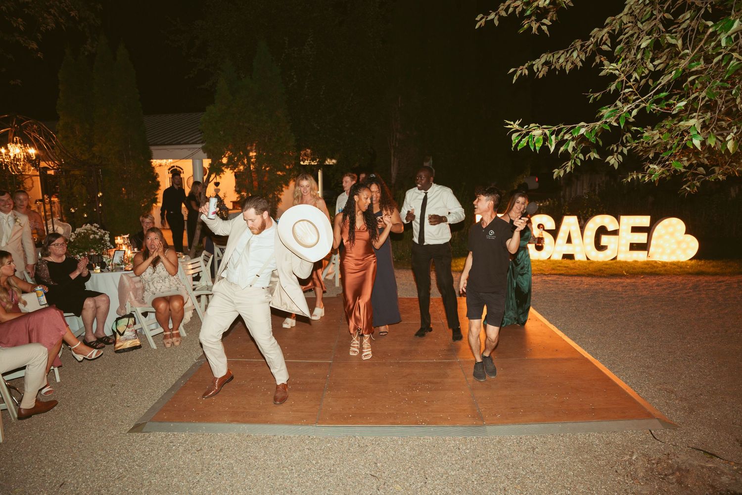 Outdoor wedding celebration at night with illuminated signage and guests dancing on patio.