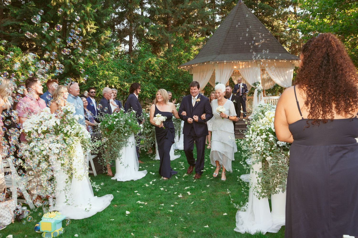 Outdoor wedding ceremony setup with white floral arrangements and guests gathering on lawn.