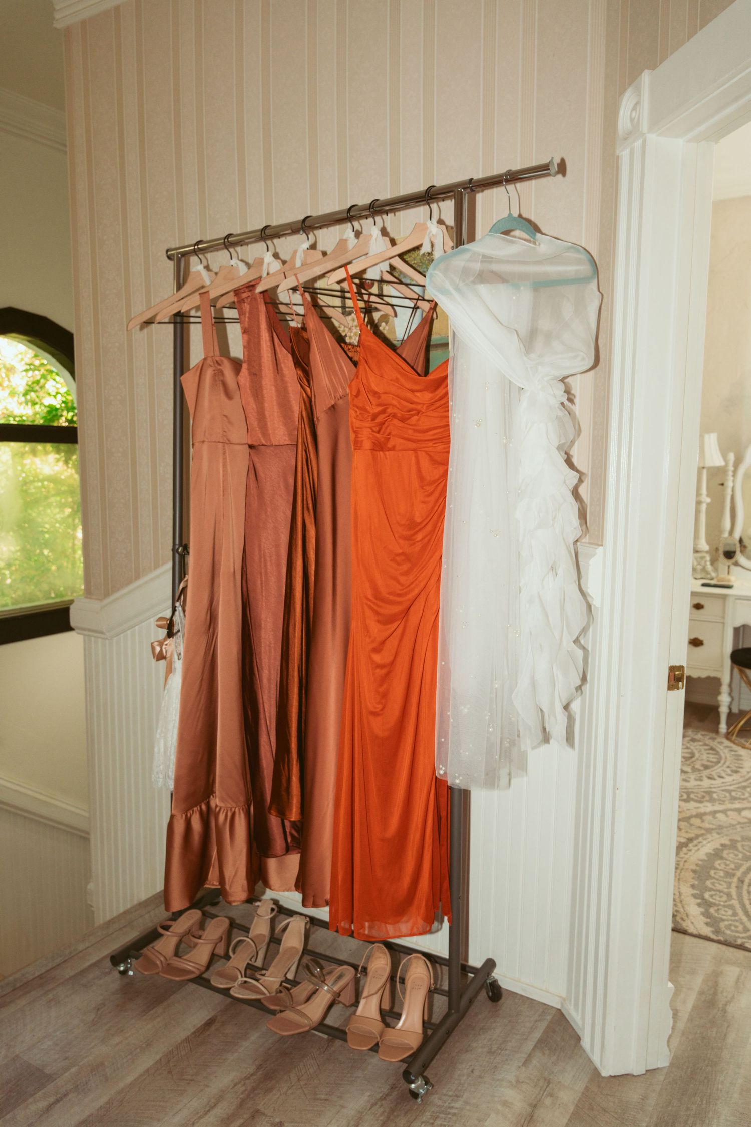 Orange and rust-colored bridesmaid dresses hanging on a rack with matching shoes below.