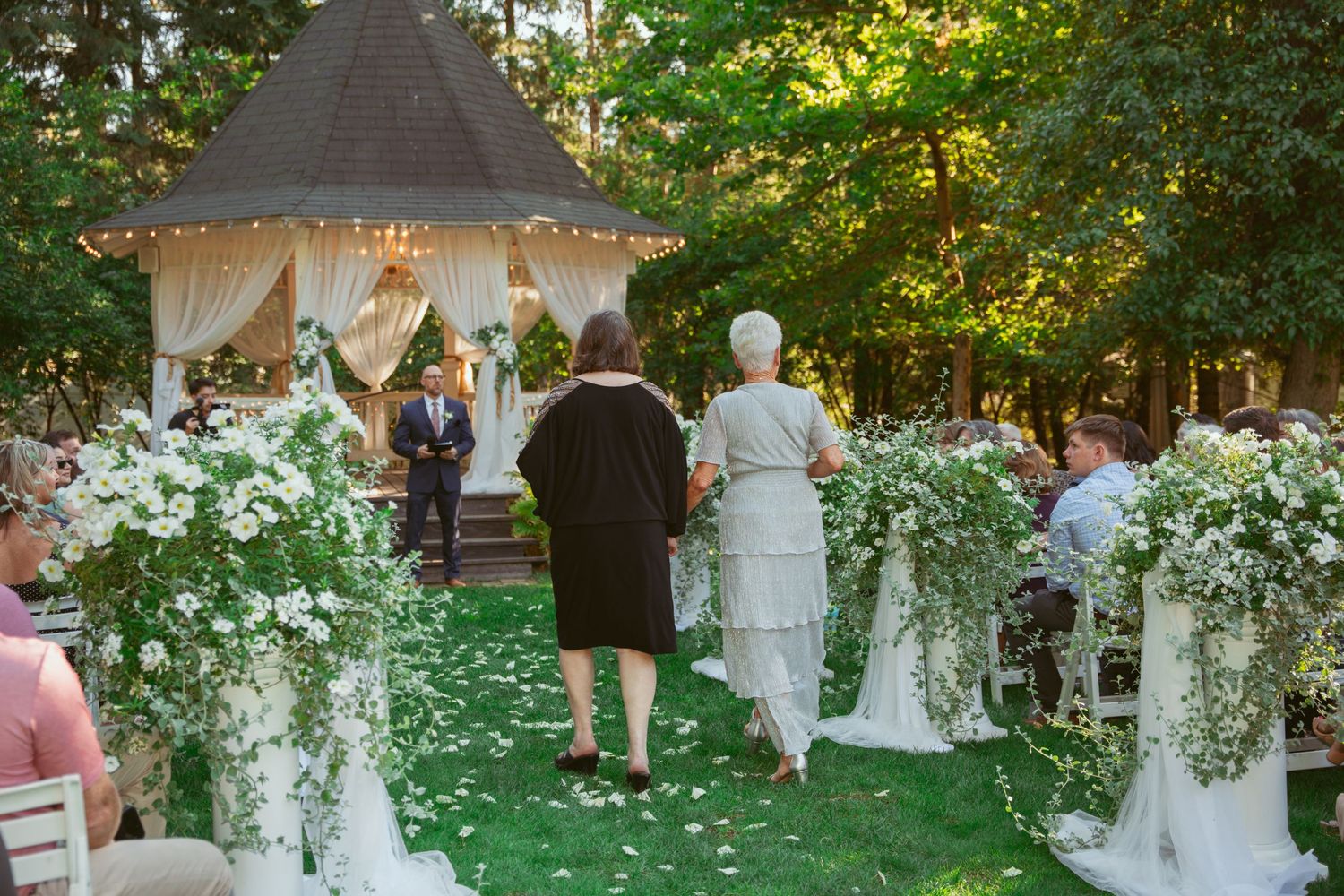 Outdoor garden wedding ceremony setup with white floral decorations and a decorated gazebo.