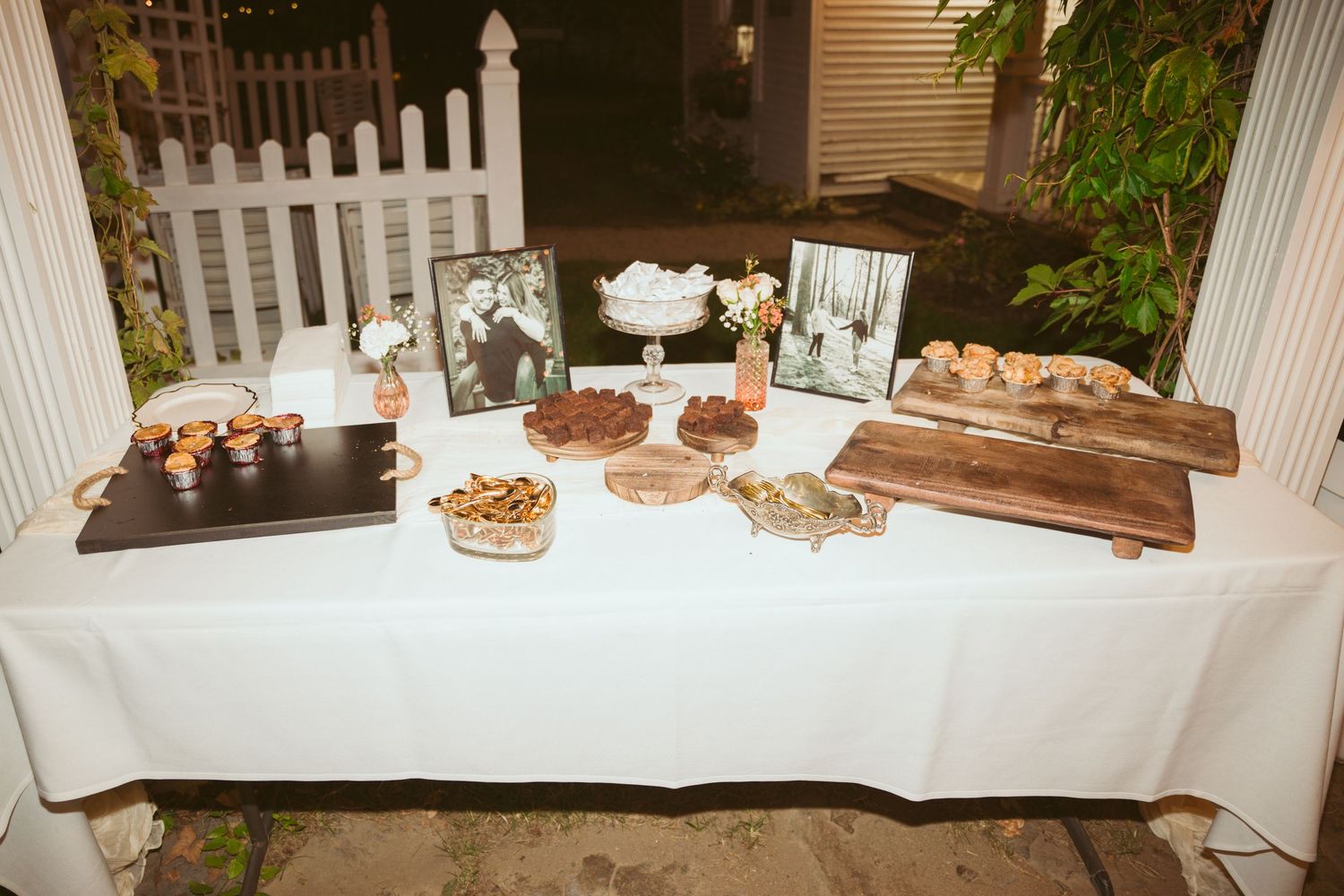 Elegant dessert table with white tablecloth displaying cakes and treats at outdoor wedding reception.