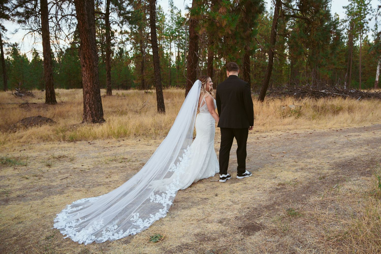 Wedding couple walking on forest path with long lace veil trailing behind.