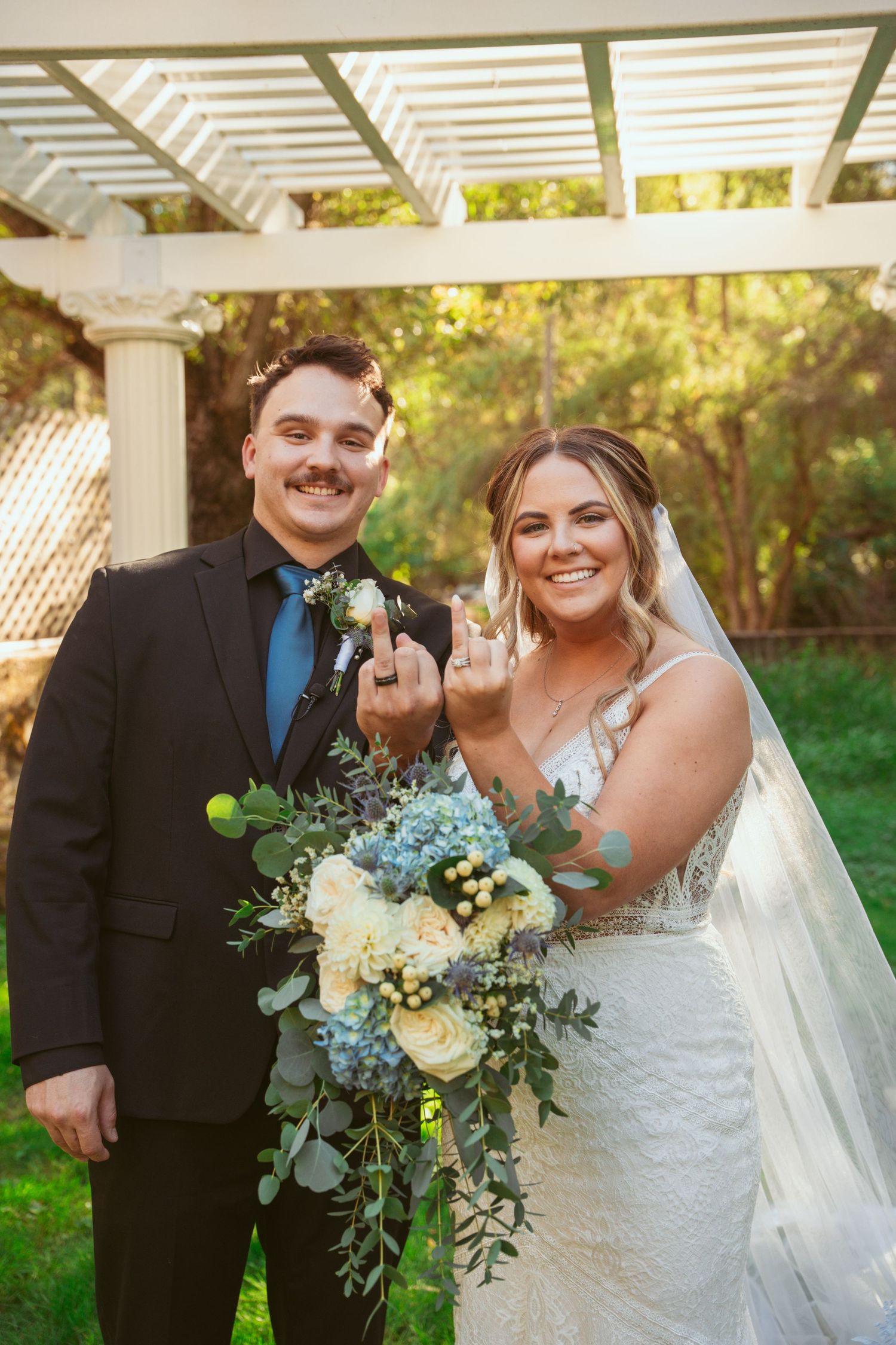 A couple celebrates their wedding day under a white pergola with blue and white floral bouquets.