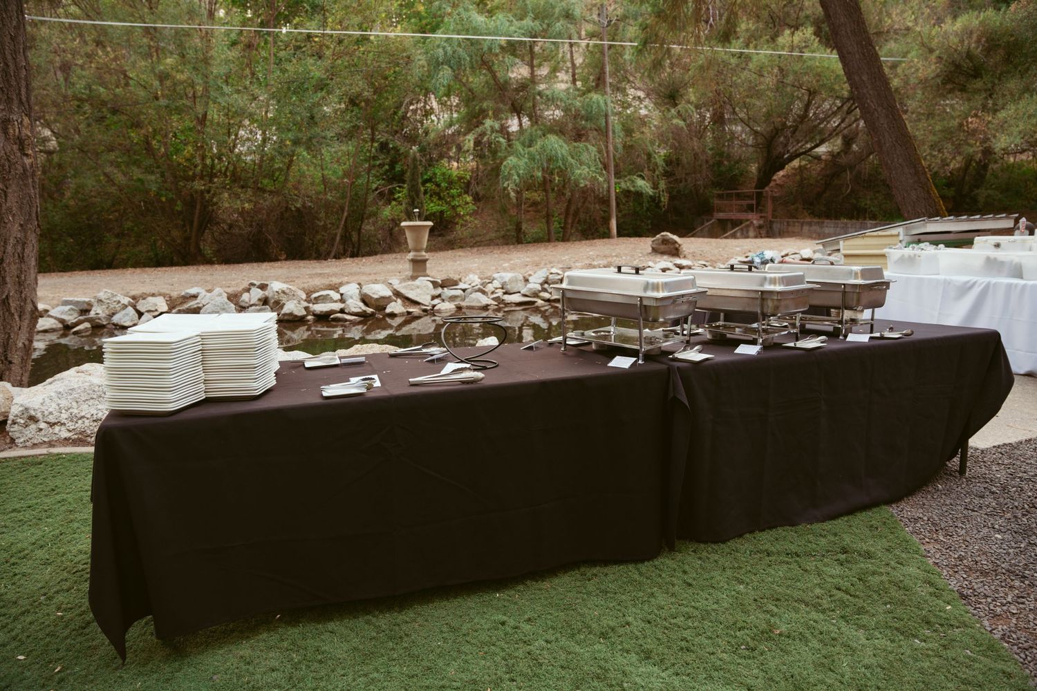 Long buffet table covered with black tablecloth and white plates set up outdoors for catering service.