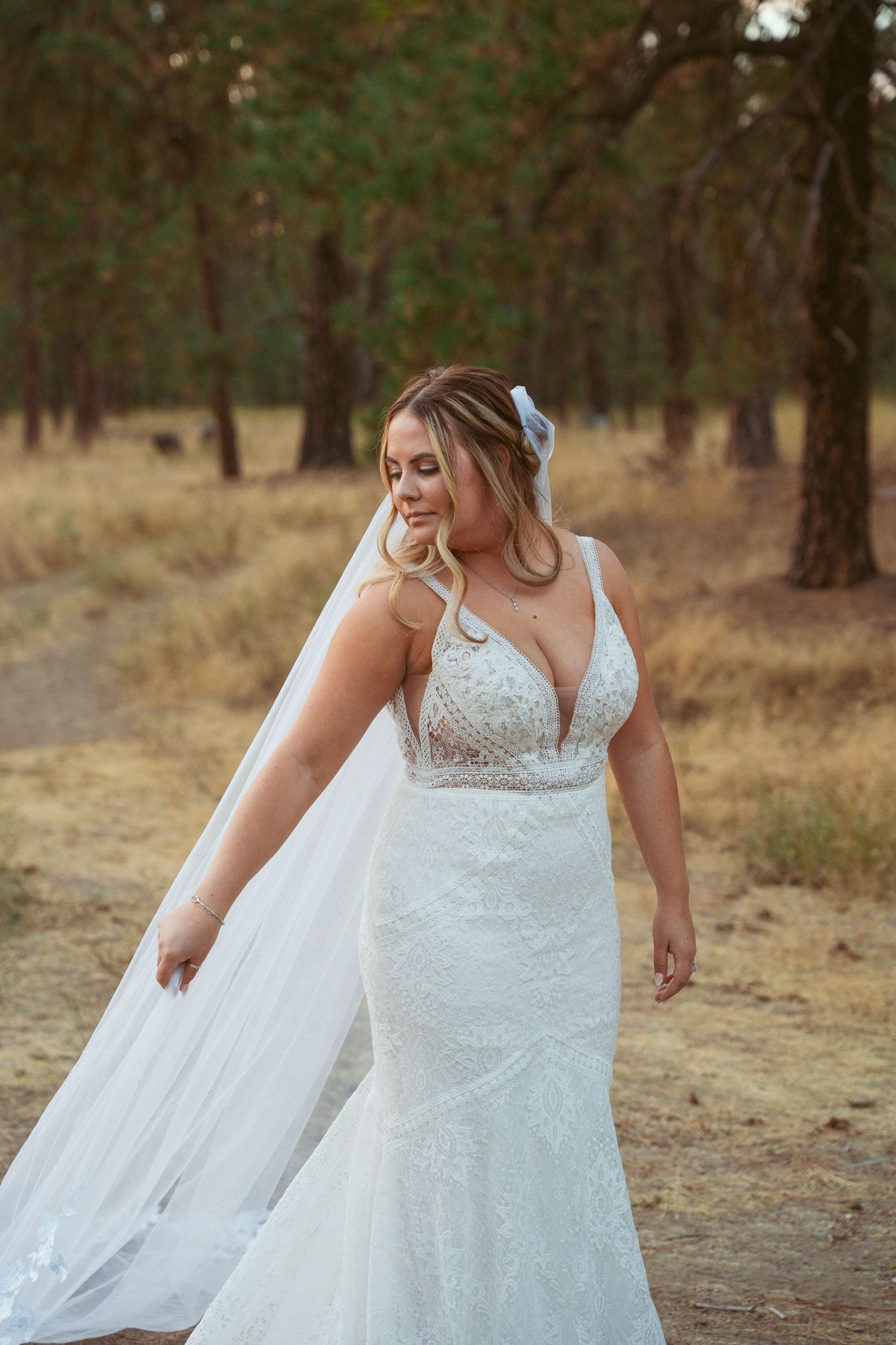 Elegant mermaid-style wedding dress with lace details and flowing veil captured in forest setting.