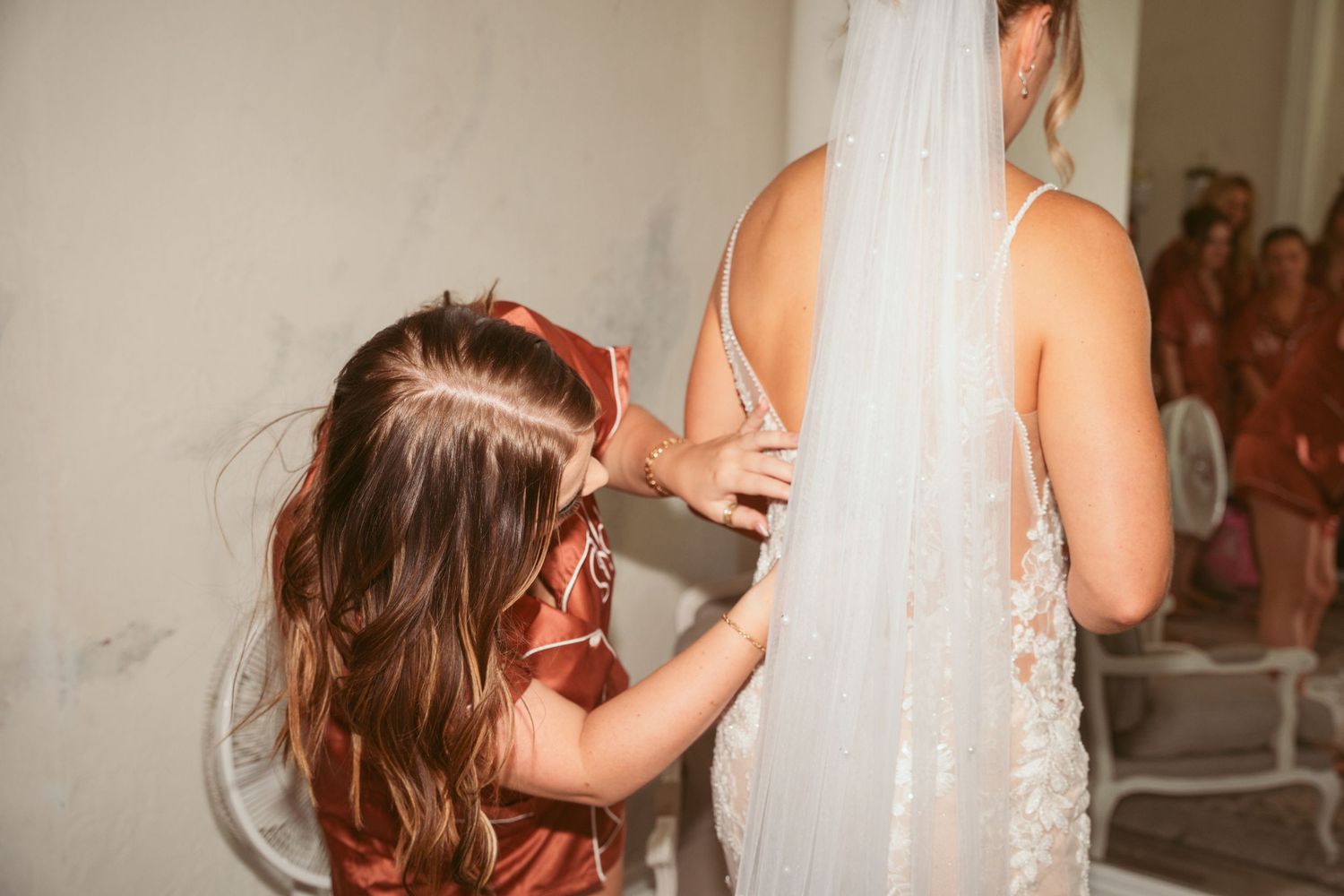 Bridesmaid in copper satin pajamas helps adjust a wedding dress during preparation.