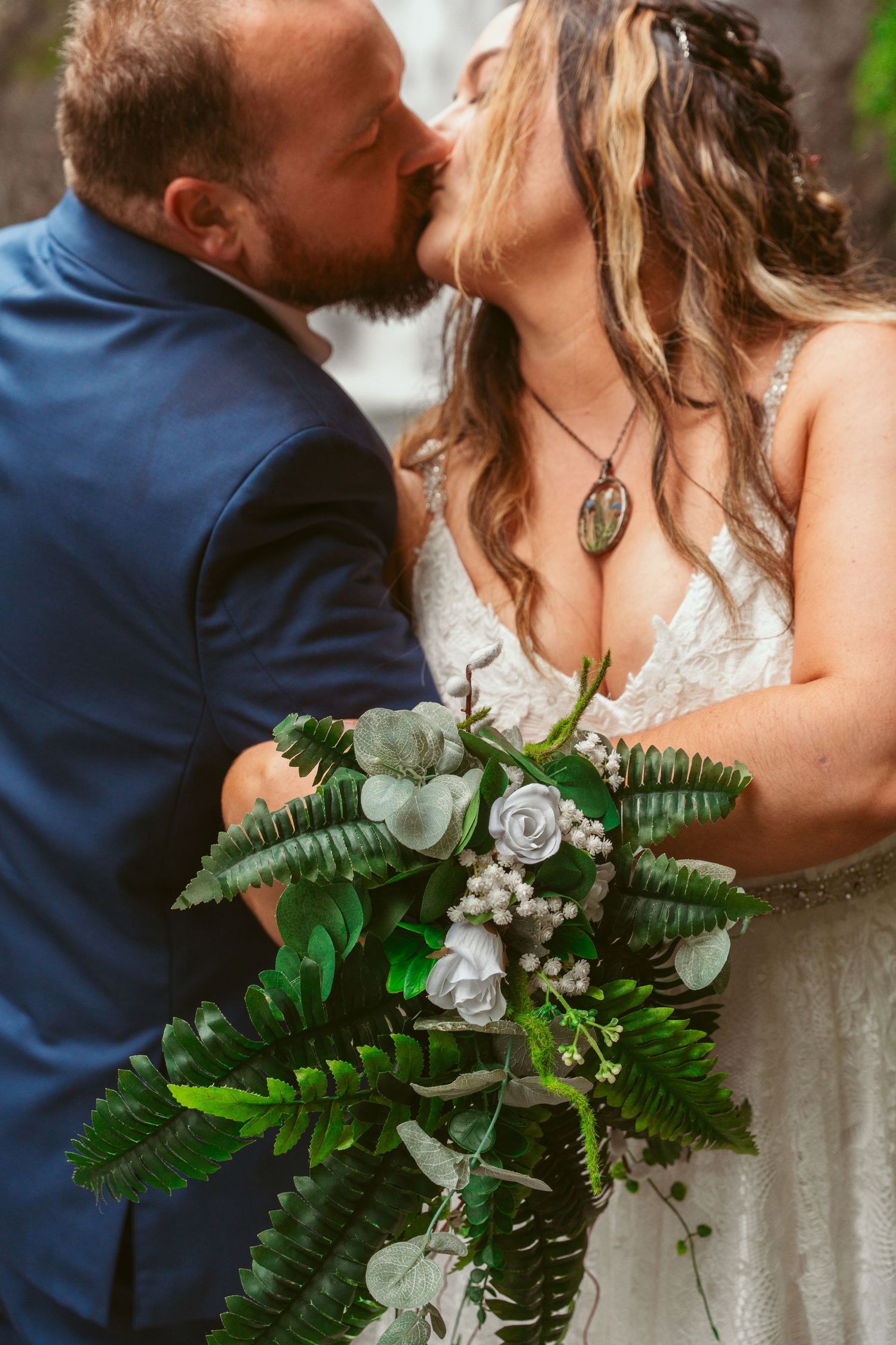 Wedding bouquet features lush green ferns and white roses against waterfall backdrop.