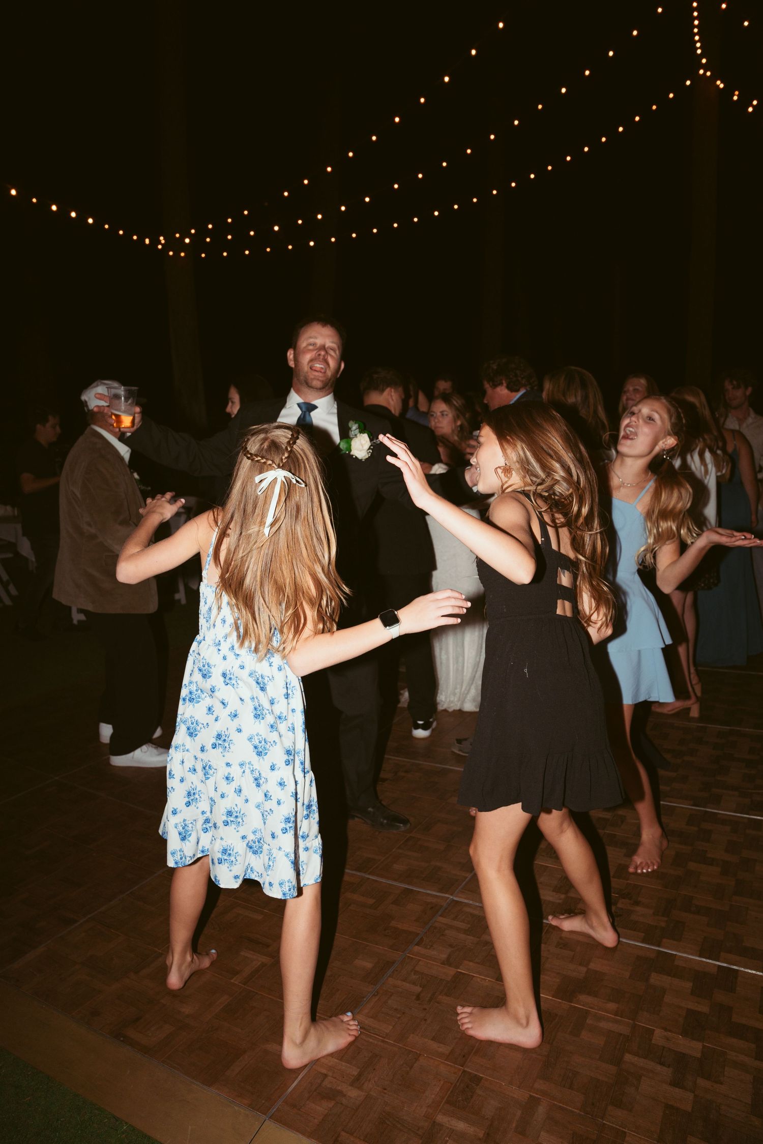 People dancing barefoot on a wooden dance floor under string lights at an evening celebration.