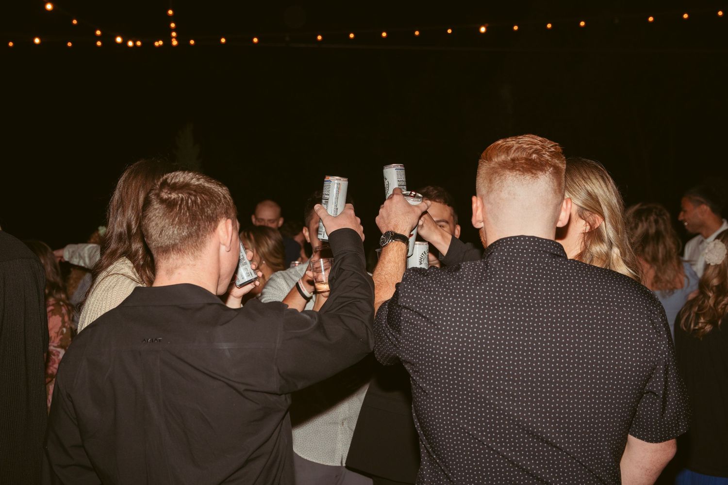 Party guests raise their drinks for a toast under string lights at a nighttime celebration.