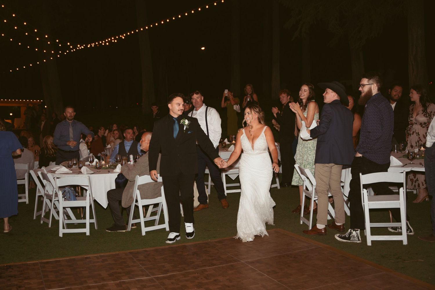 String lights illuminate an outdoor nighttime wedding reception with guests dancing.