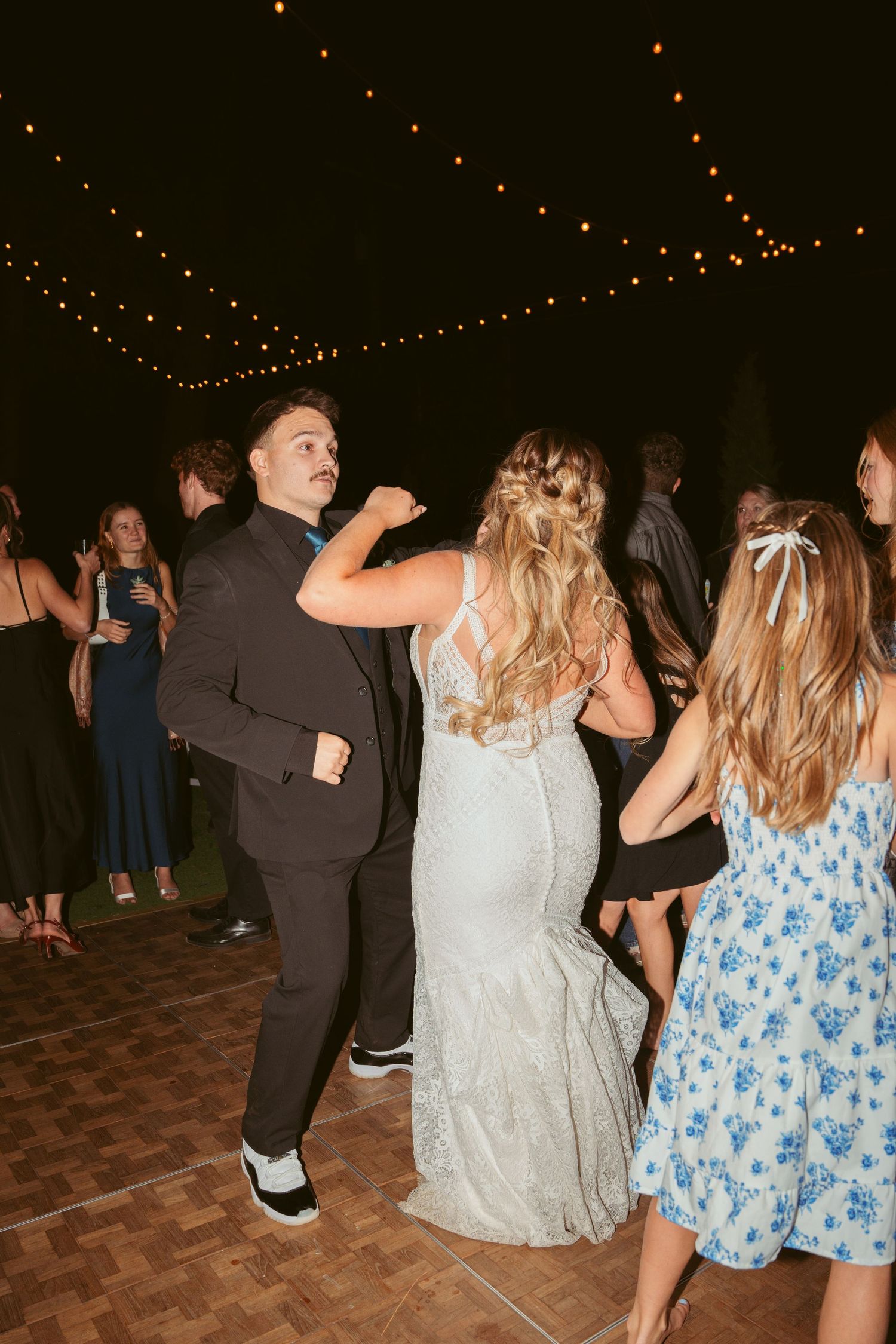 People dancing at a wedding reception under decorative string lights on a wooden dance floor.