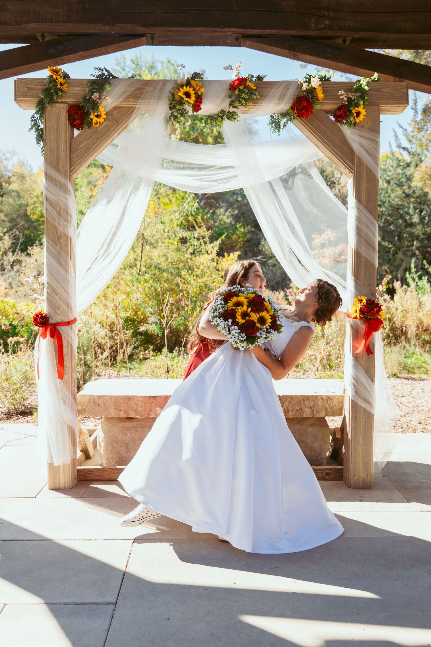 Wedding celebration captured under wooden pergola with elegant white draping and sunflower decorations.