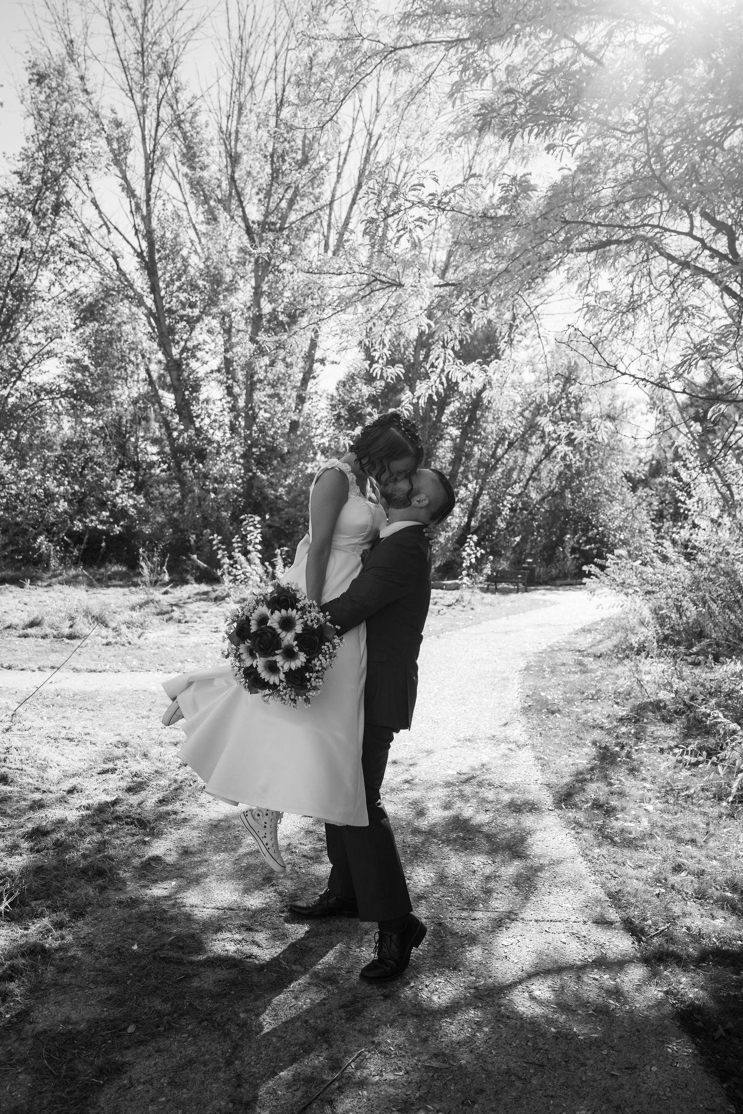A black and white wedding photo showing a romantic embrace on a sunlit forest path.