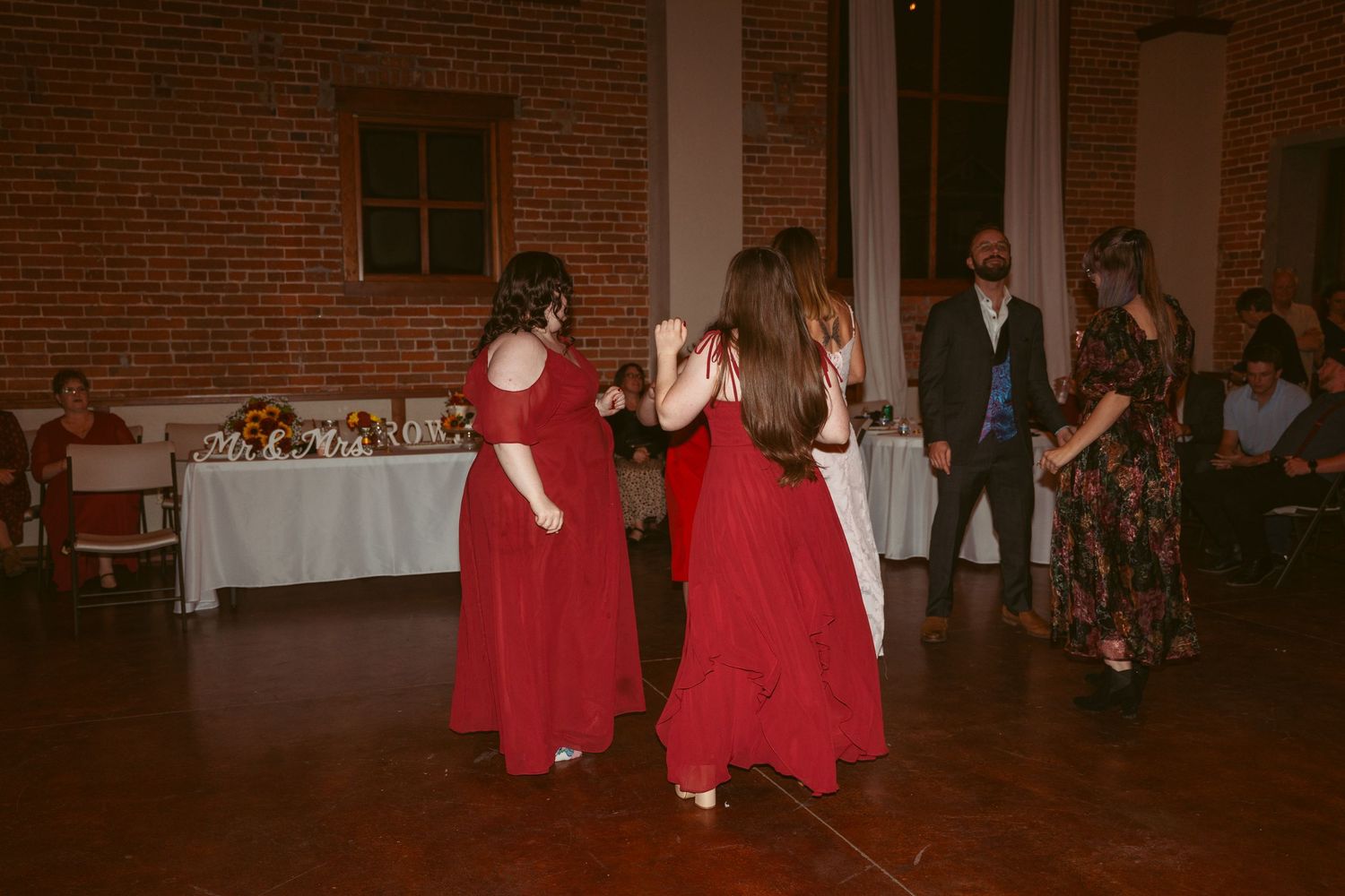 People dancing in formal attire under string lights in a brick-walled reception venue.