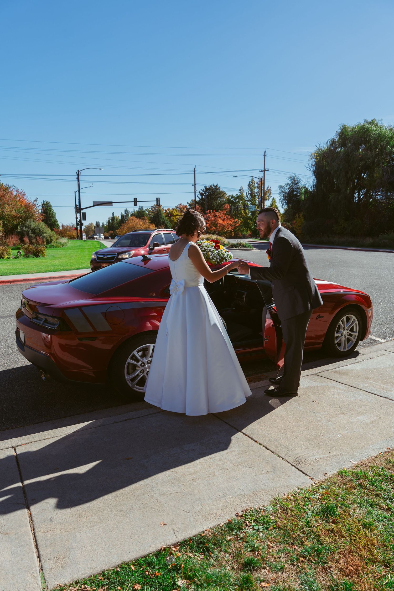 Wedding couple poses next to their red car on a beautiful autumn day.