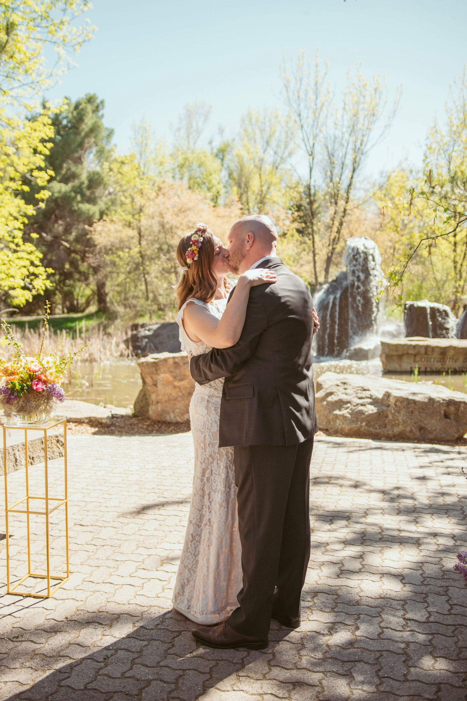 A romantic wedding couple embraces by an ornate stone fountain in a garden setting.