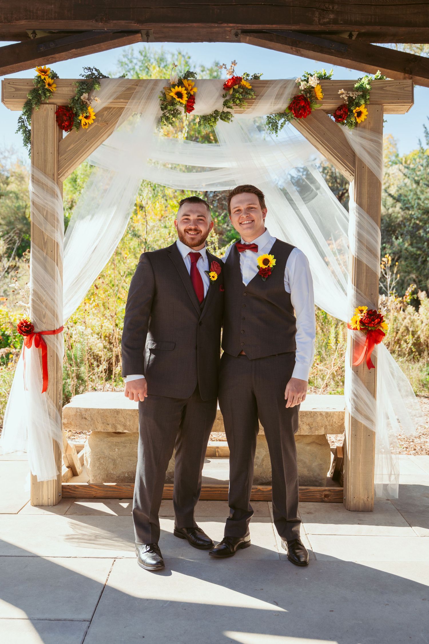 Wedding party members pose together under rustic pavilion with autumn floral decorations.