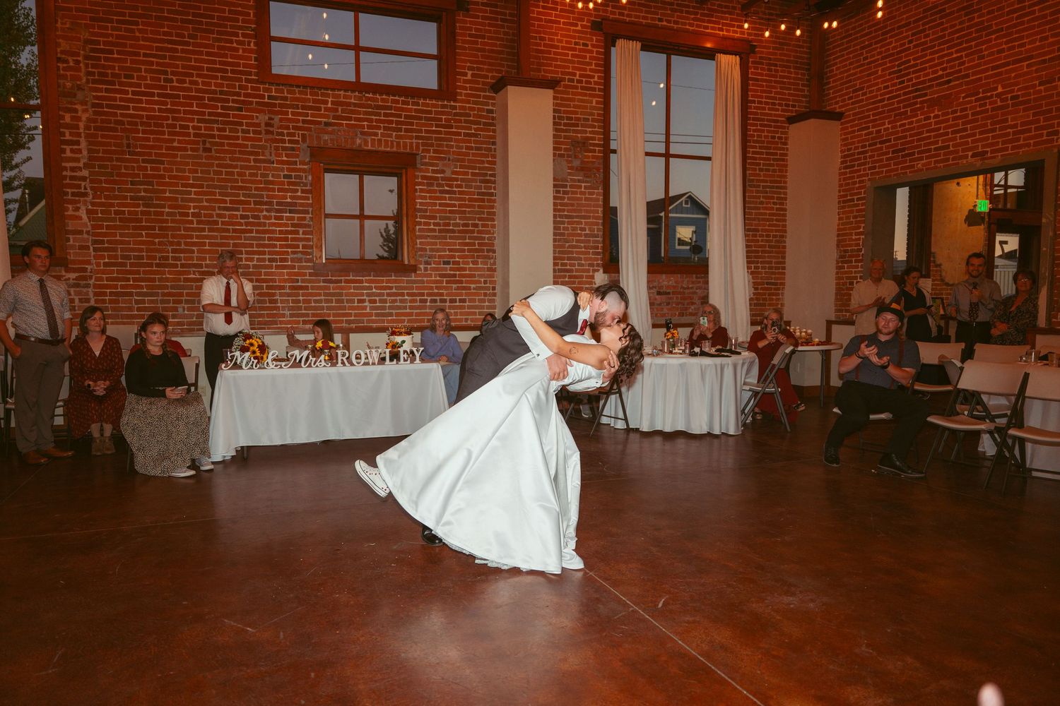 Wedding dancers execute a dramatic dip move in a rustic venue with exposed brick walls.