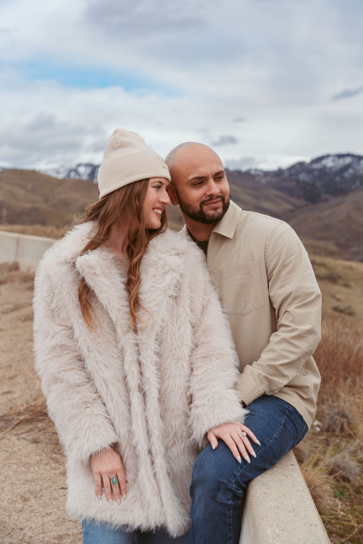 A couple in warm winter outfits sharing a tender moment against a backdrop of snow-capped mountains on a cloudy day.