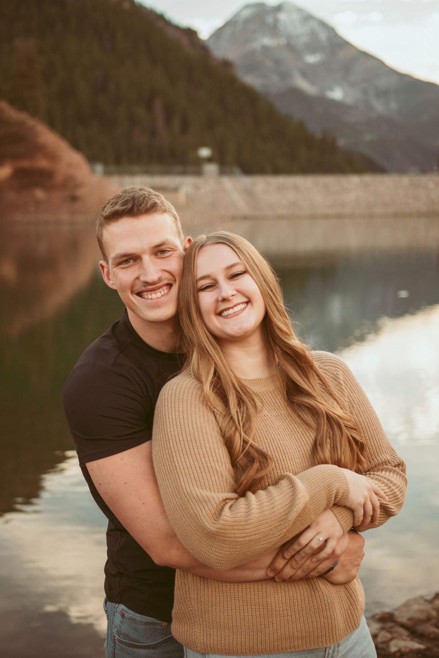 Couple embracing and laughing by a scenic mountain lake during sunset.