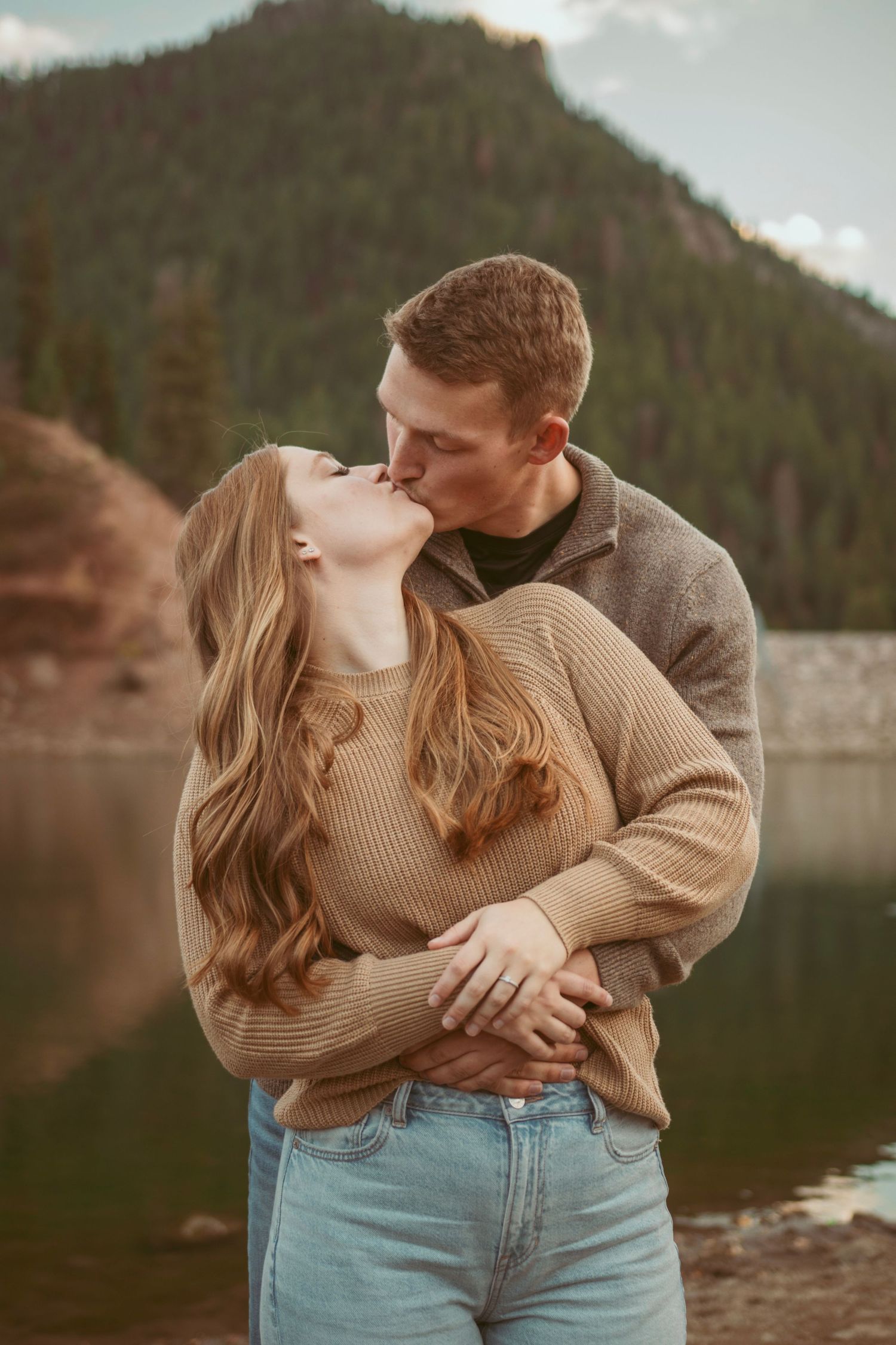 A romantic embrace by a mountain lake with two people wearing sweaters and jeans.