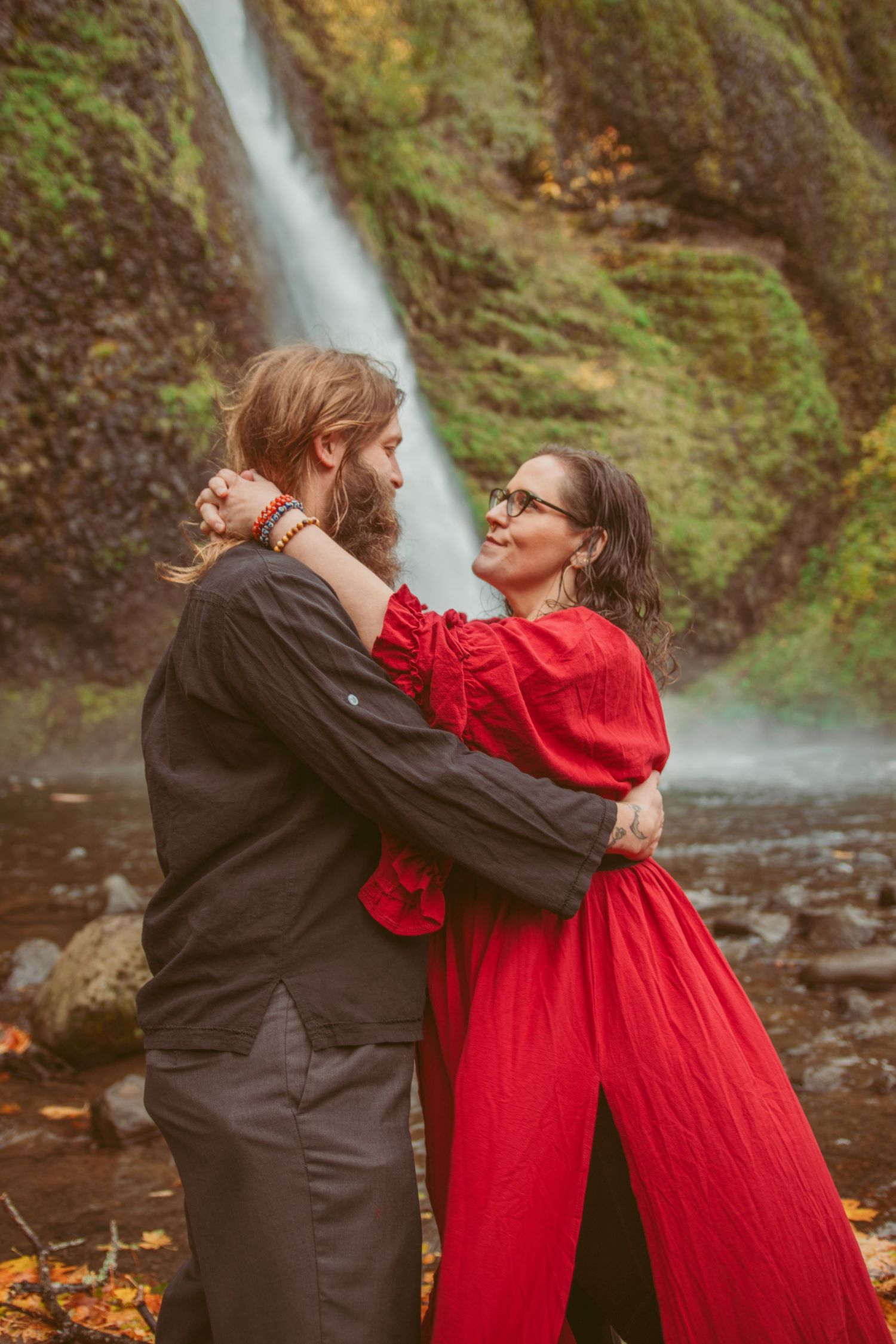 A couple in contrasting red and black outfits dance together in front of a cascading waterfall.