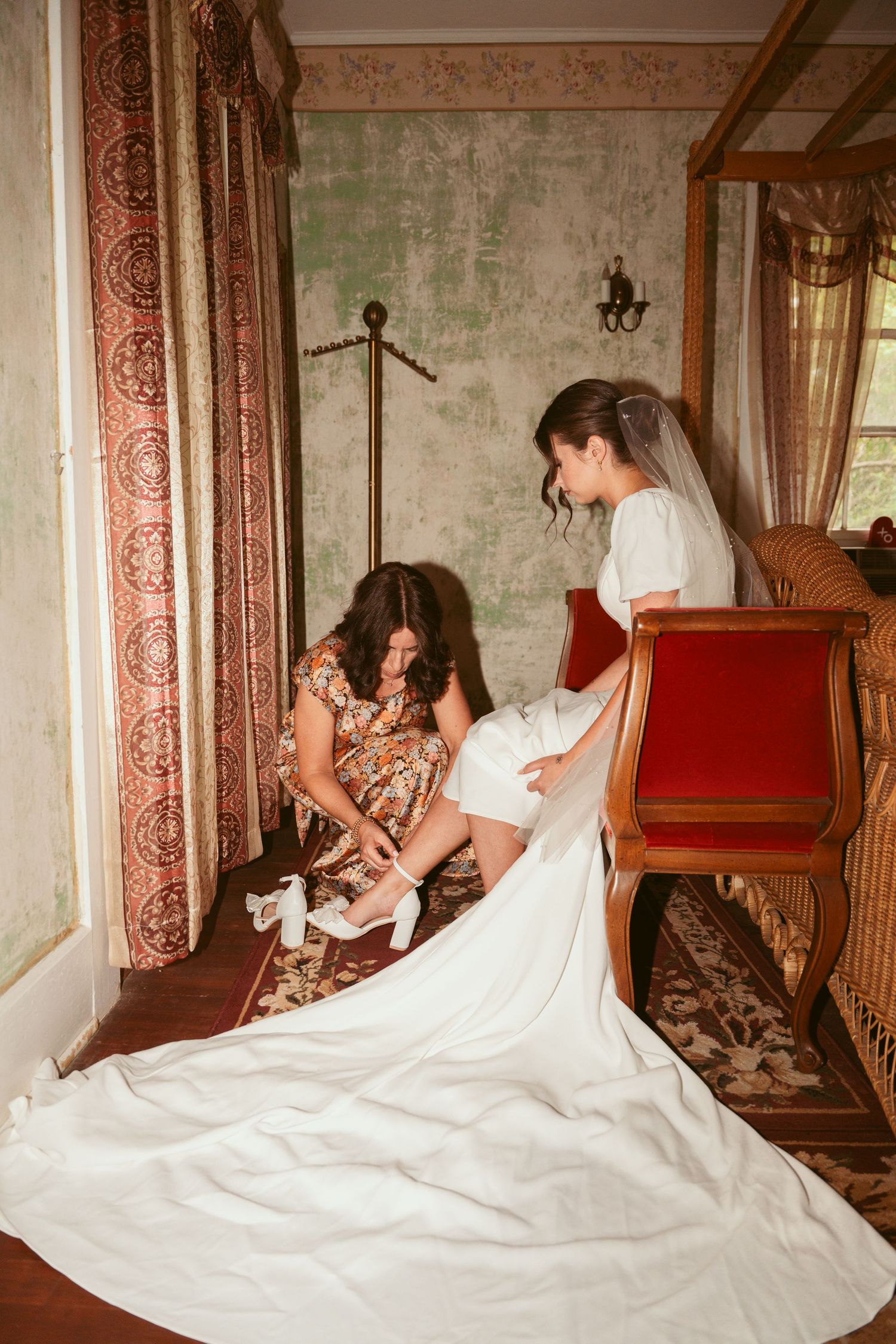 Wedding dress preparation scene on vintage stairs with floral curtains and antique decor.