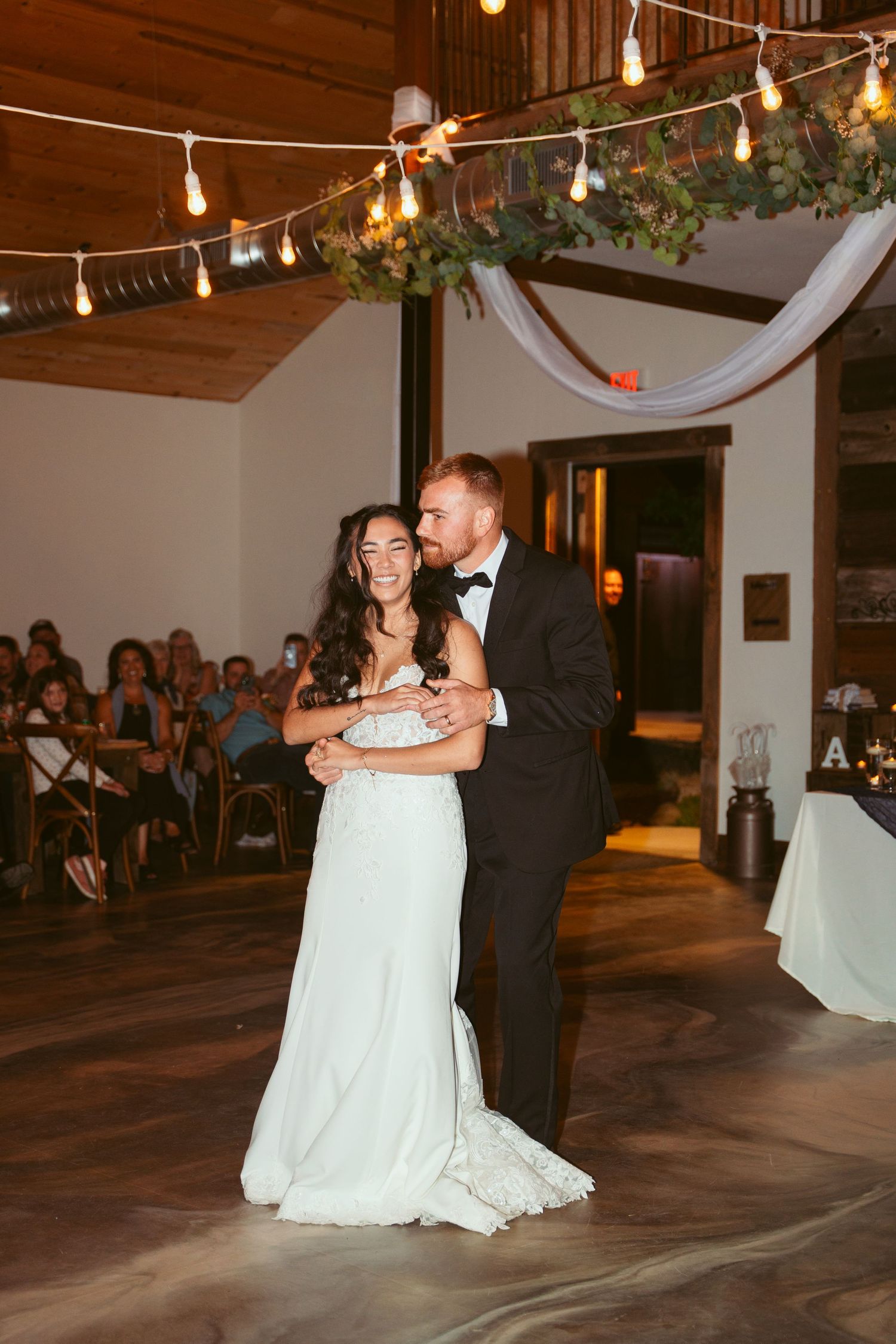 Couple sharing their first dance under string lights in a rustic wedding venue.