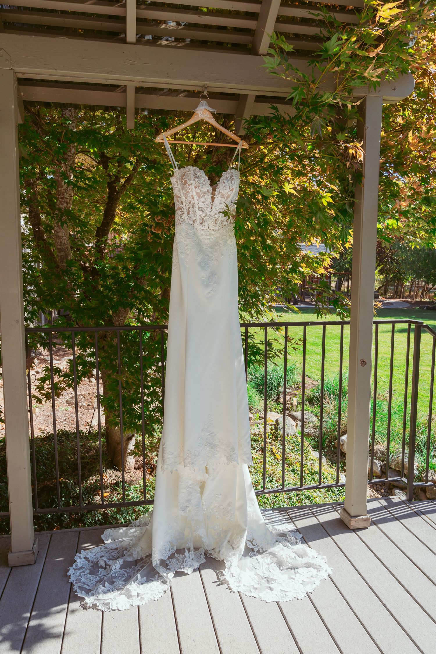 Elegant white lace wedding dress displayed on covered outdoor porch with garden view.
