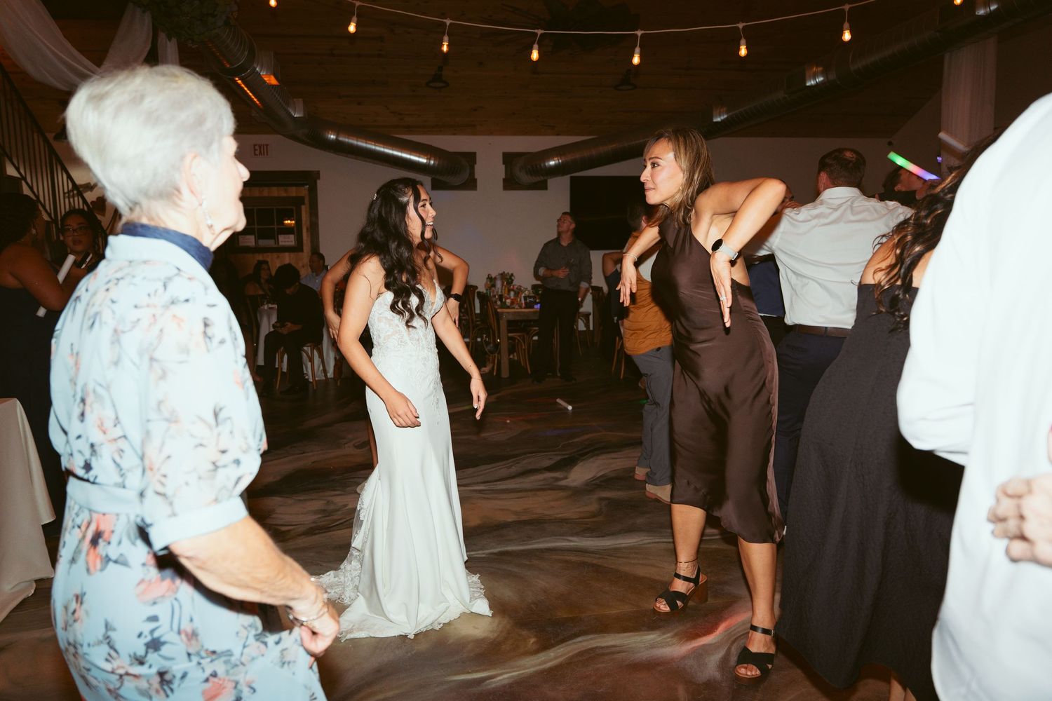 Wedding reception guests dancing under warm string lights in an indoor venue.