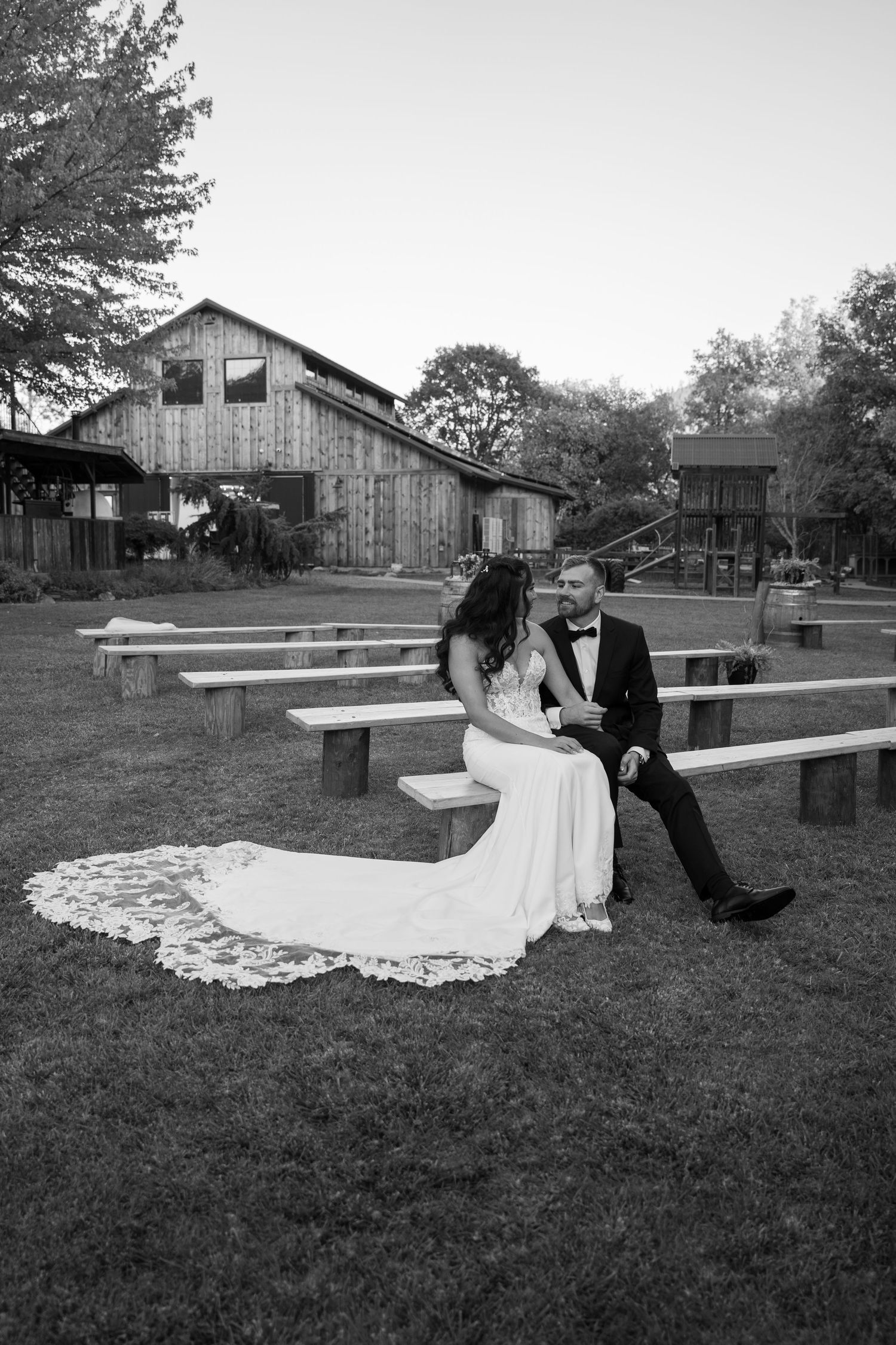 Black and white panorama of newlyweds sitting on benches with rustic buildings in background.