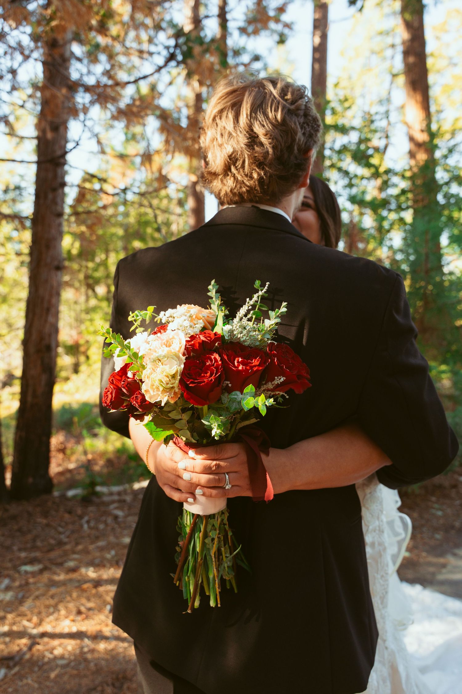 A wedding couple embracing while holding a vibrant red and white bouquet in a forest.