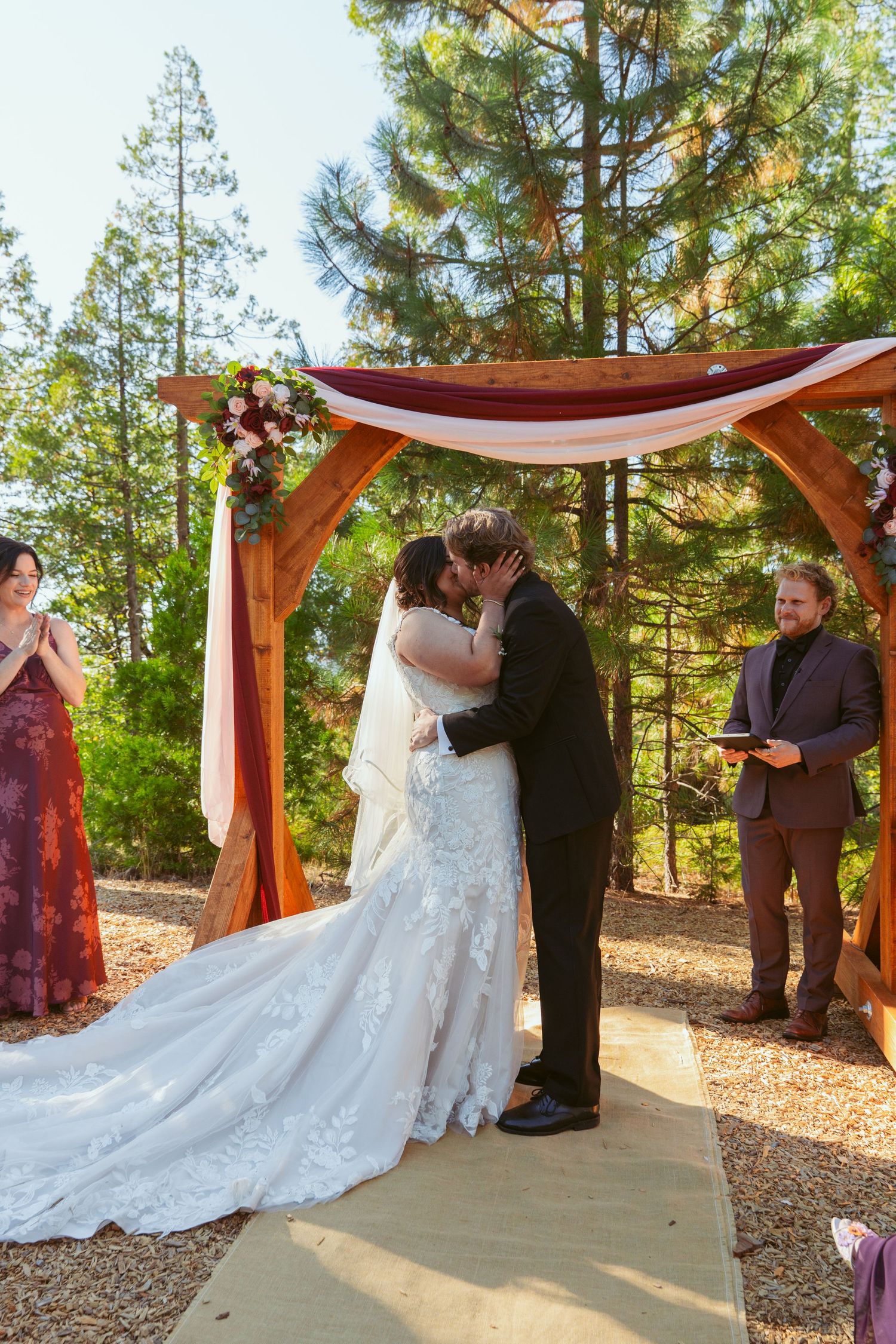 Wedding ceremony under wooden arch with white draping and floral arrangements in a forest setting.