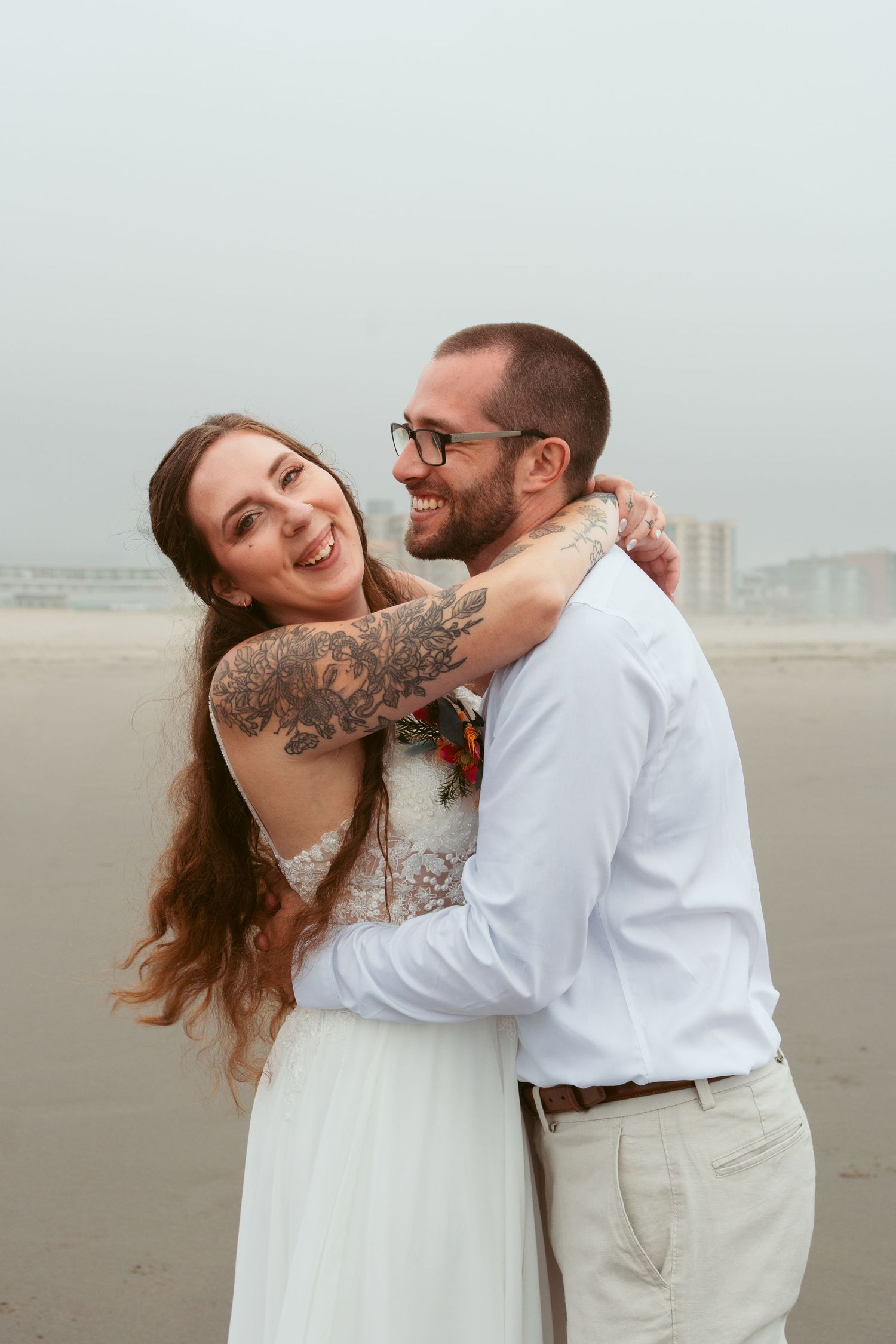 A couple laughs together during their beach wedding photoshoot with cloudy skies in the background.