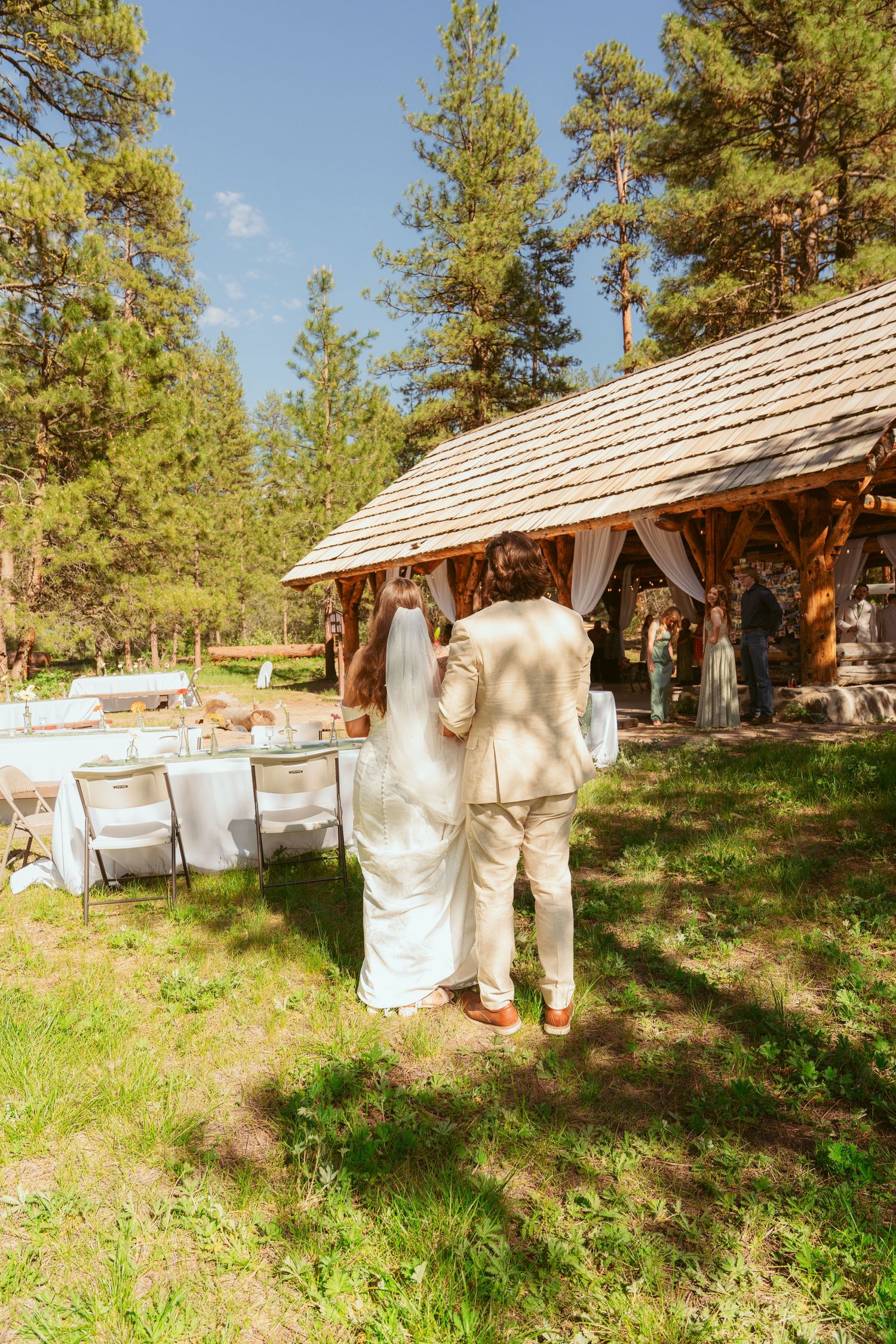 A rustic outdoor wedding reception setup with a wooden pavilion and white tables in a forest setting.