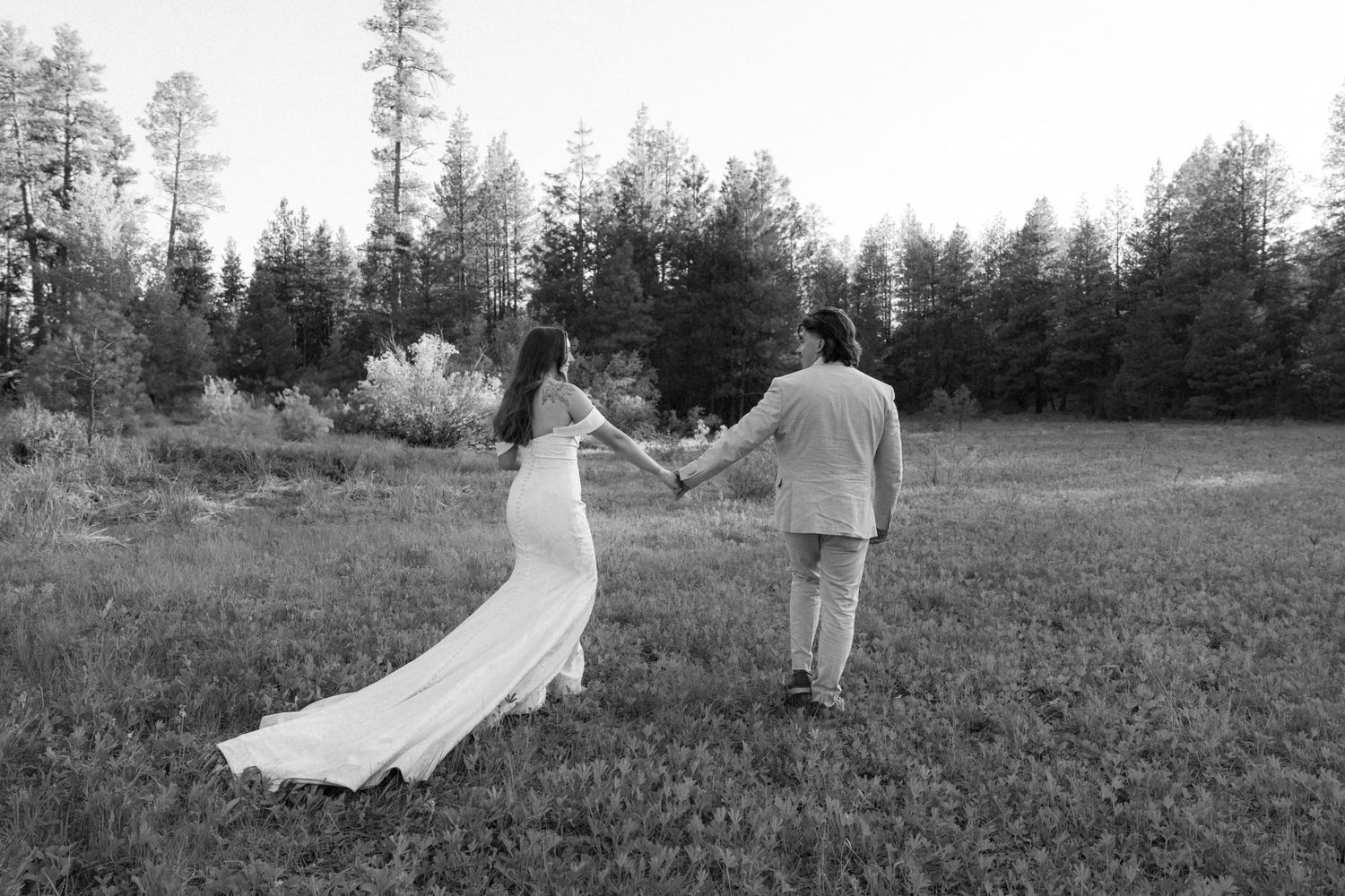 Black and white photo of newlyweds dancing in an open field with pine trees on the horizon.