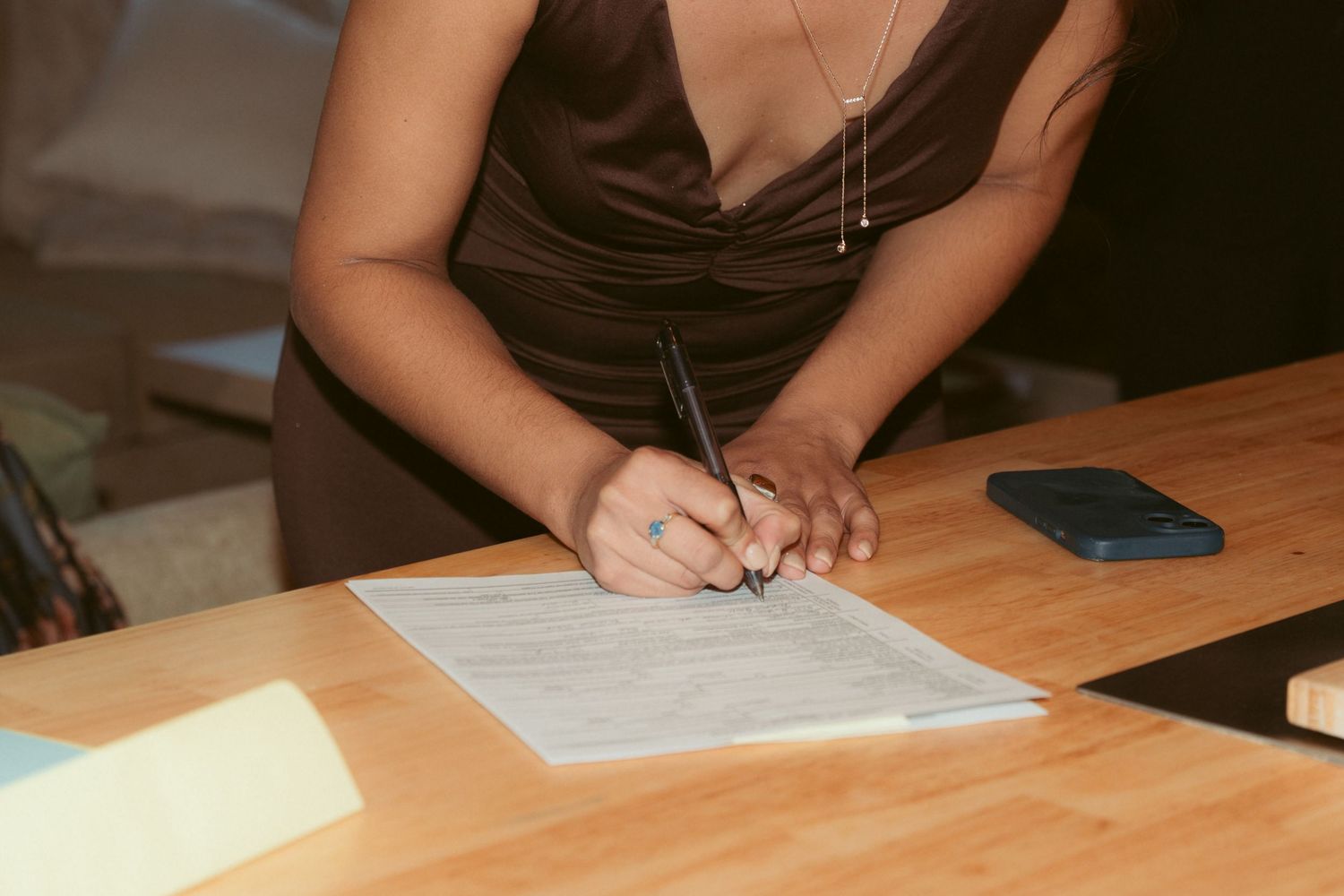 Person in brown dress signing documents at wooden table with phone nearby.