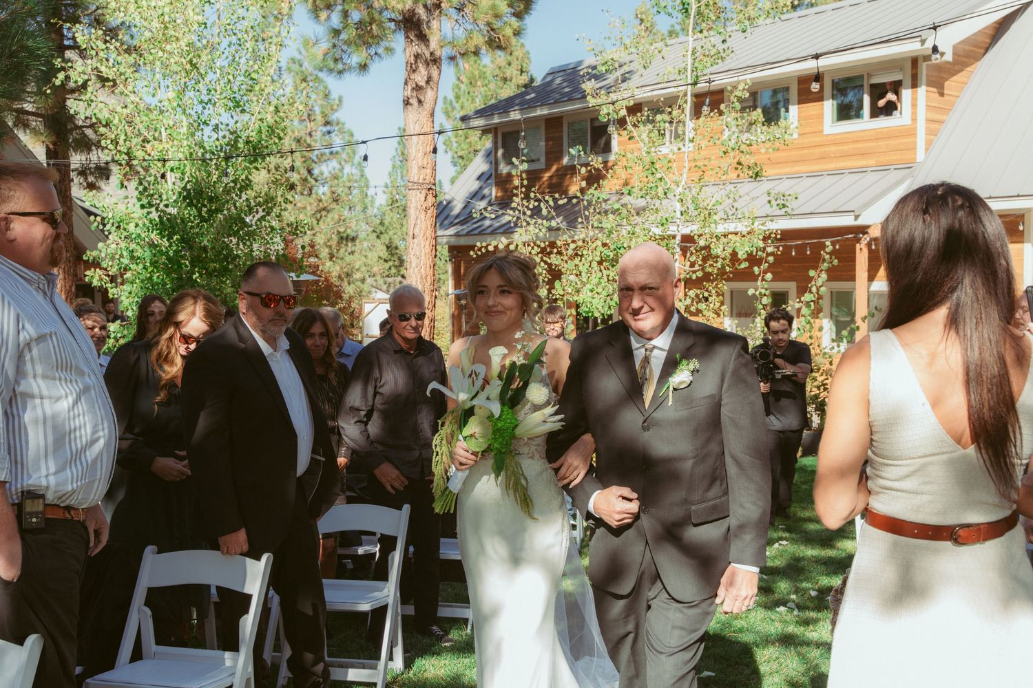 Garden wedding ceremony with guests seated on white chairs surrounded by trees.