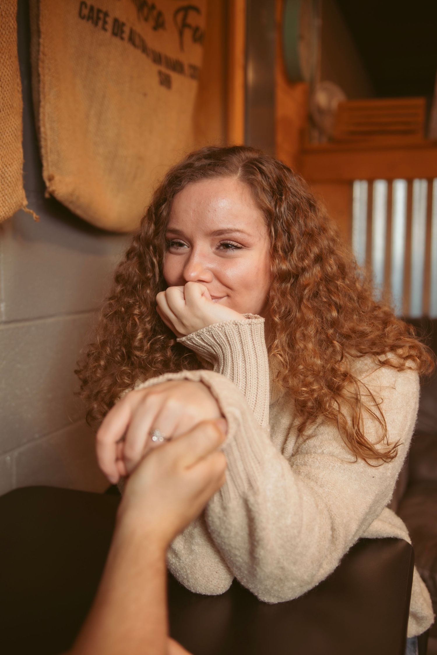 Person in cream sweater laughing with hands covering face in cafe setting.
