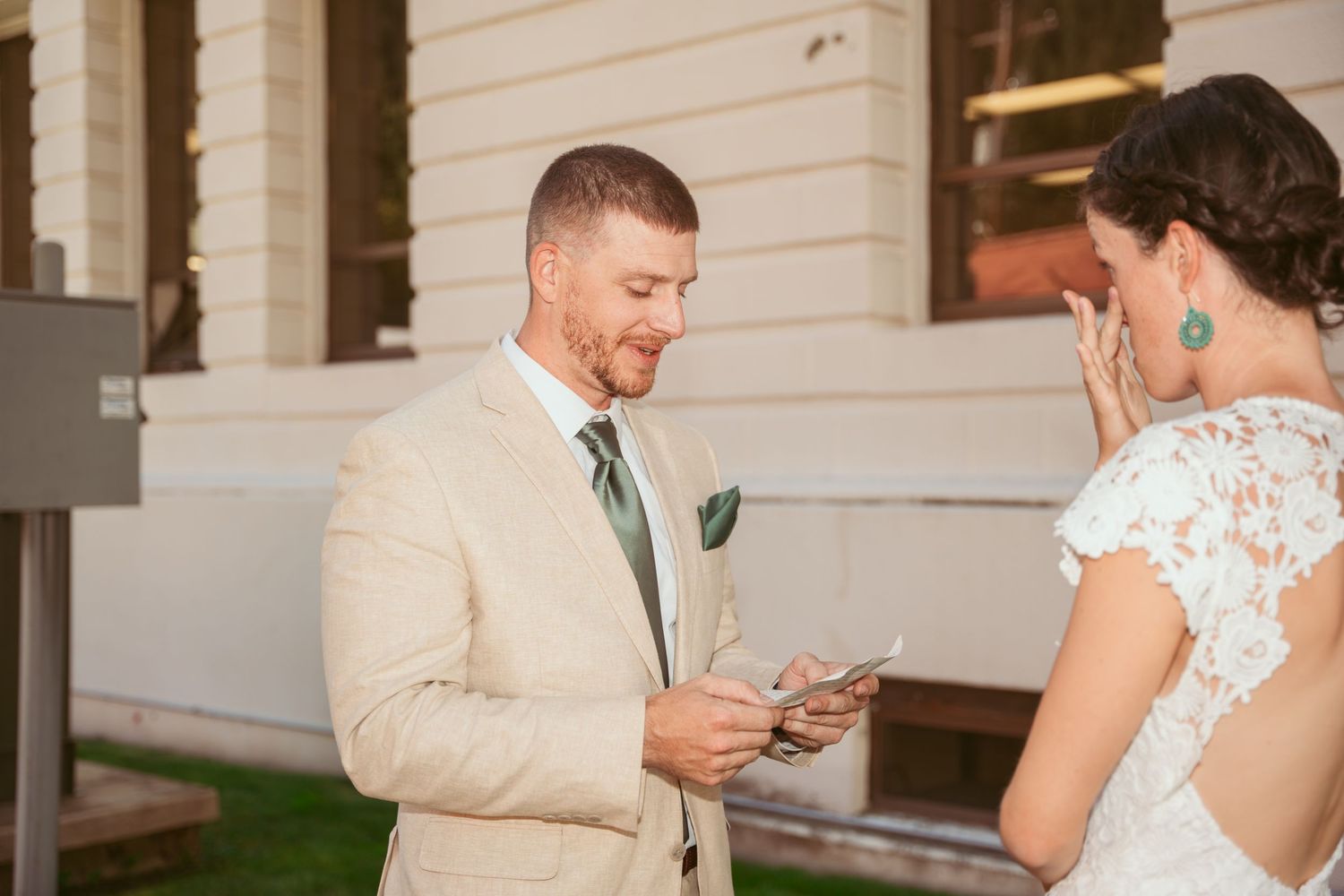 Two people in formal attire exchange notes while standing outside a white building.