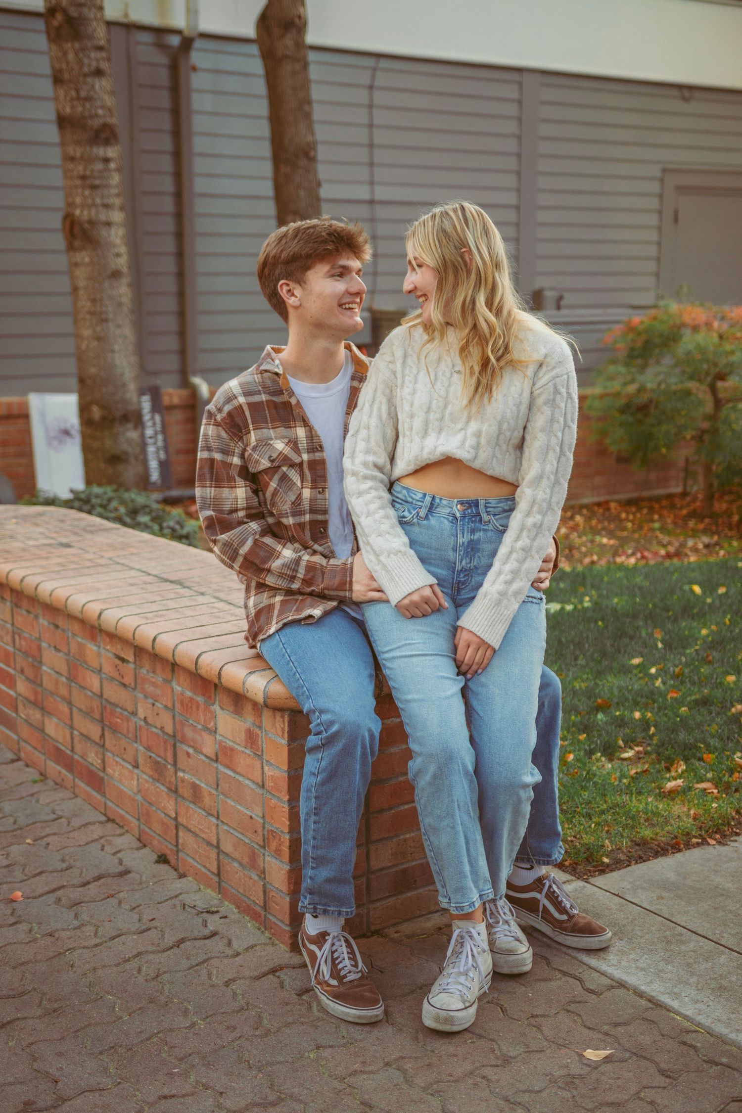 A couple in casual fall outfits sit together on a brick wall during golden hour.