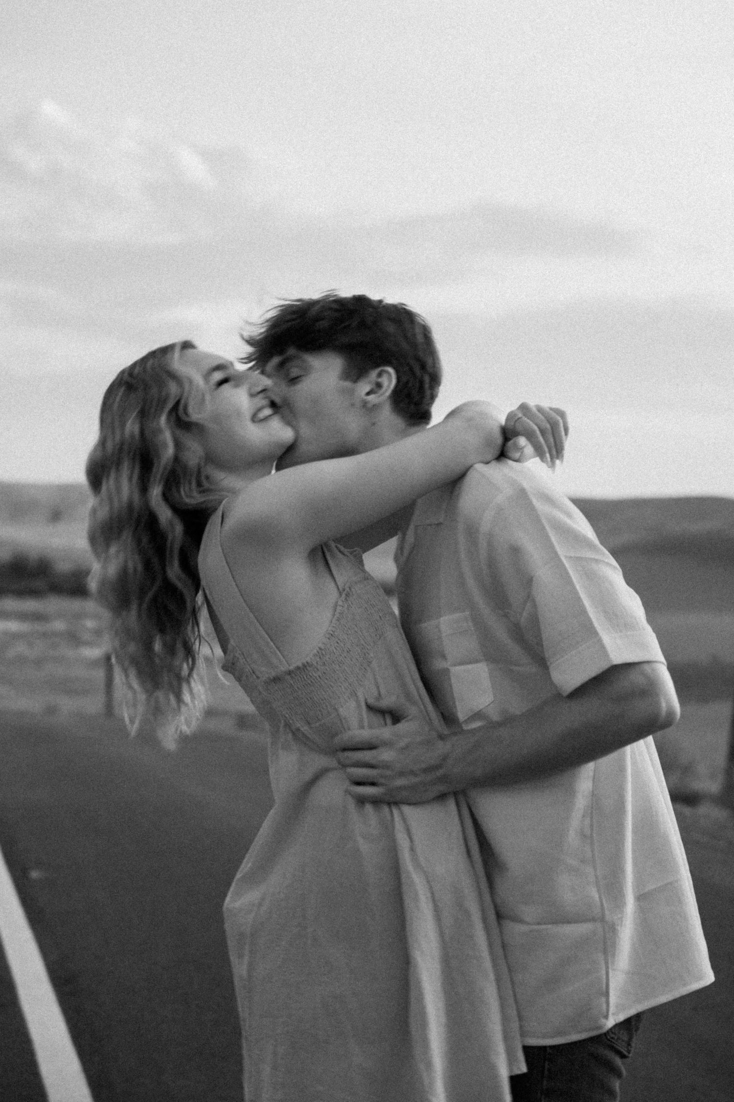 A romantic couple embraces and kisses on an empty road at sunset with mountains in the background.