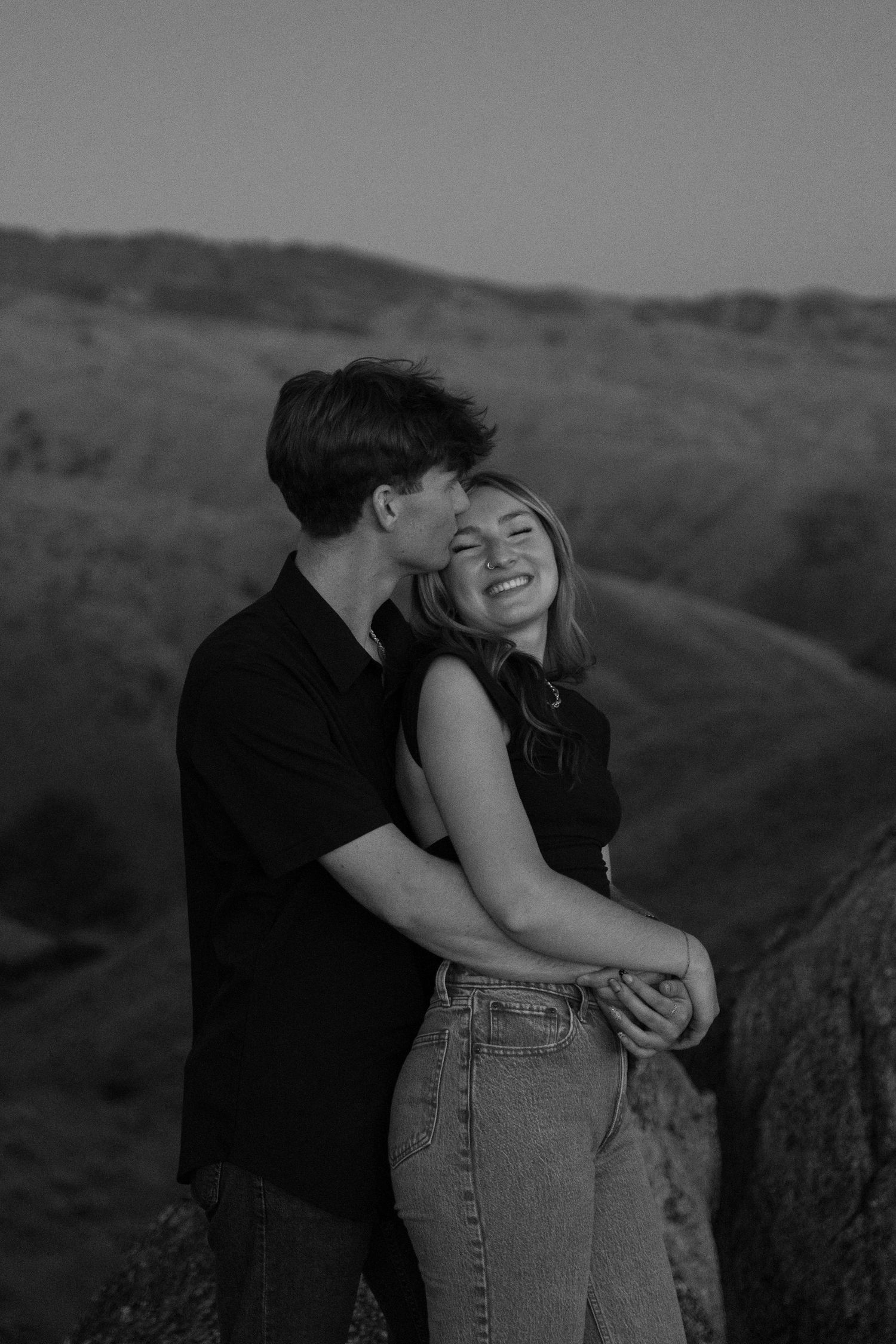 Black and white portrait of couple embracing on hilltop during sunset photoshoot.