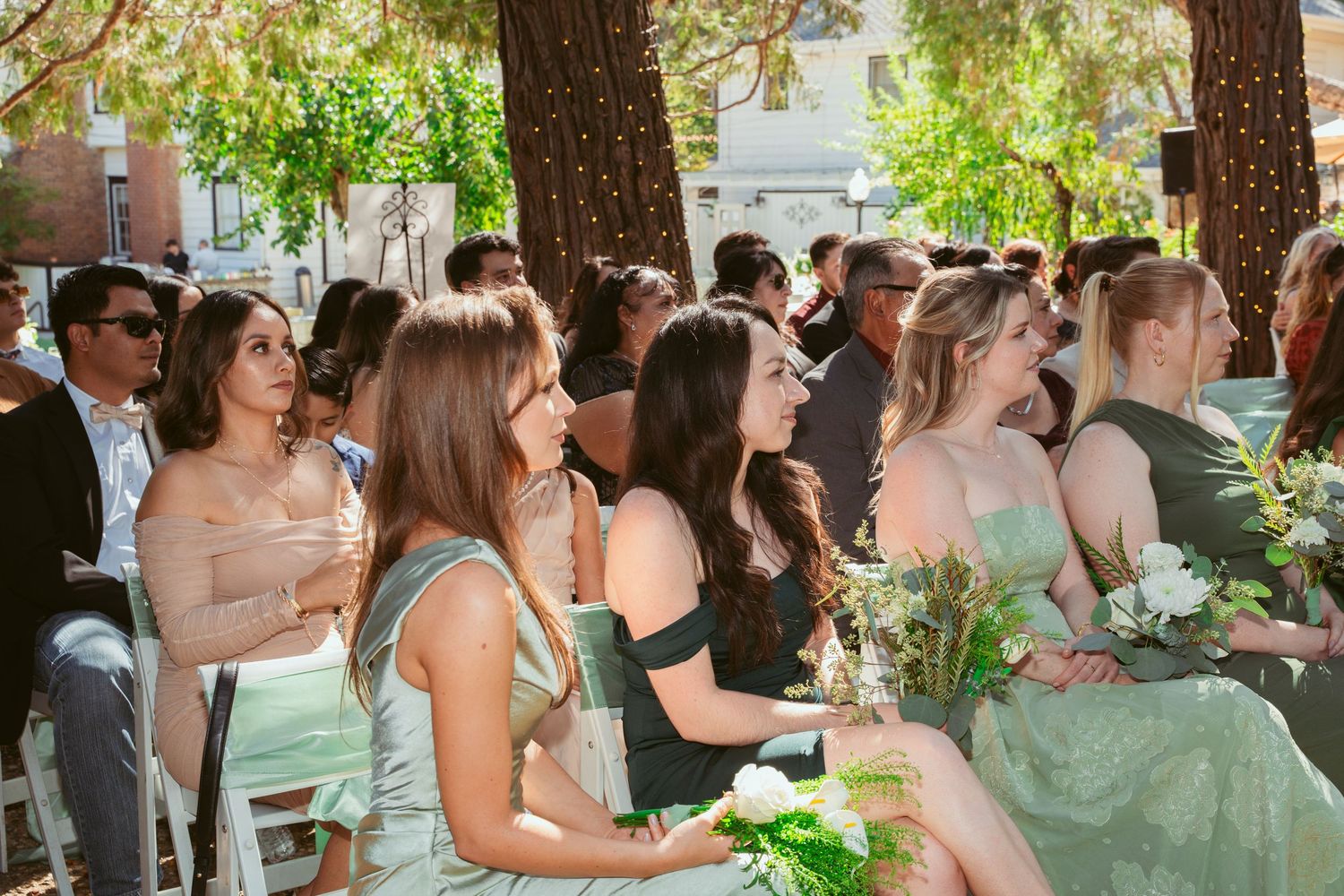 Wedding guests seated in white chairs during outdoor ceremony under lush green trees.