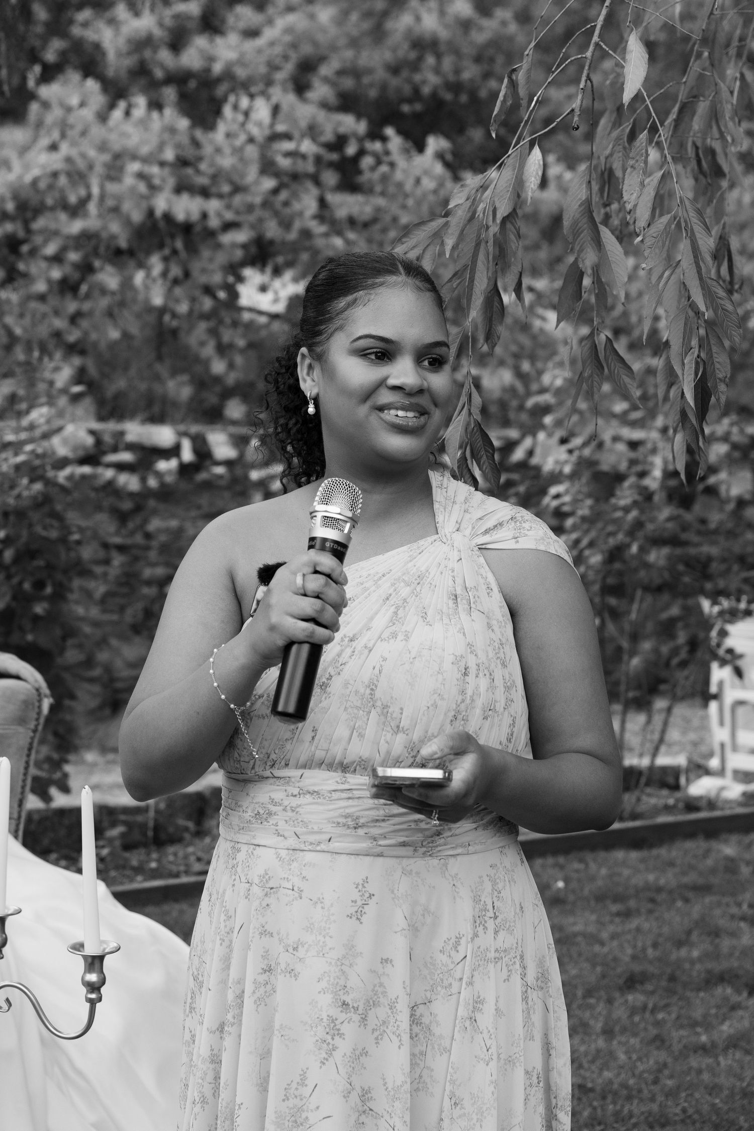 A speaker in an elegant white dress holds a microphone during an outdoor event.