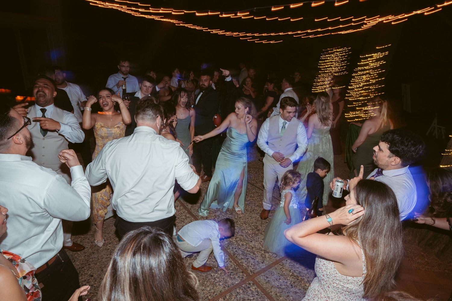 Energetic crowd dancing and celebrating at a nighttime wedding reception under string lights.