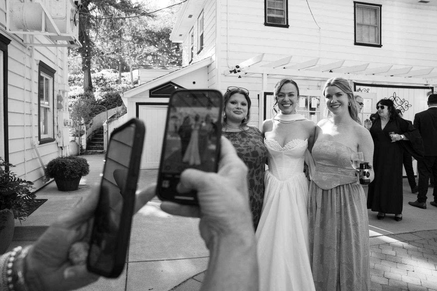 Wedding guests take photos of the bridal party outside a white house.