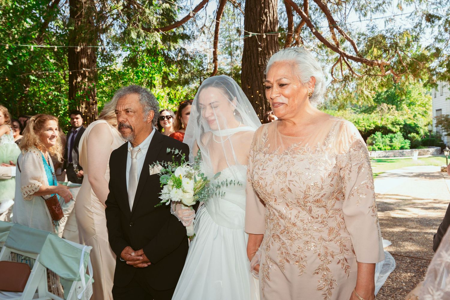 Wedding guests gather outdoors under trees for a joyful celebration in a garden setting.