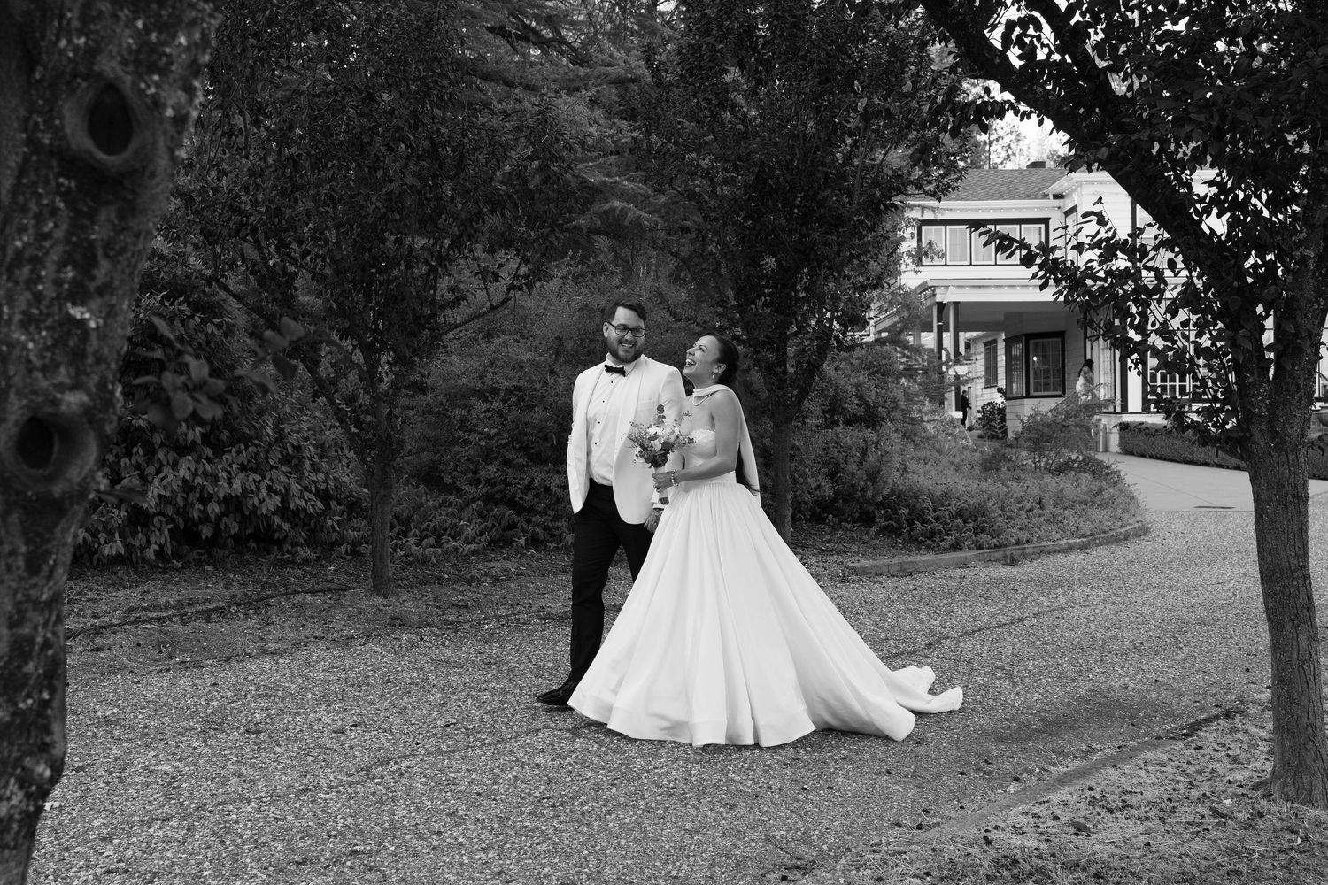 A romantic wedding couple poses for photos in black and white on a gravel path surrounded by trees.