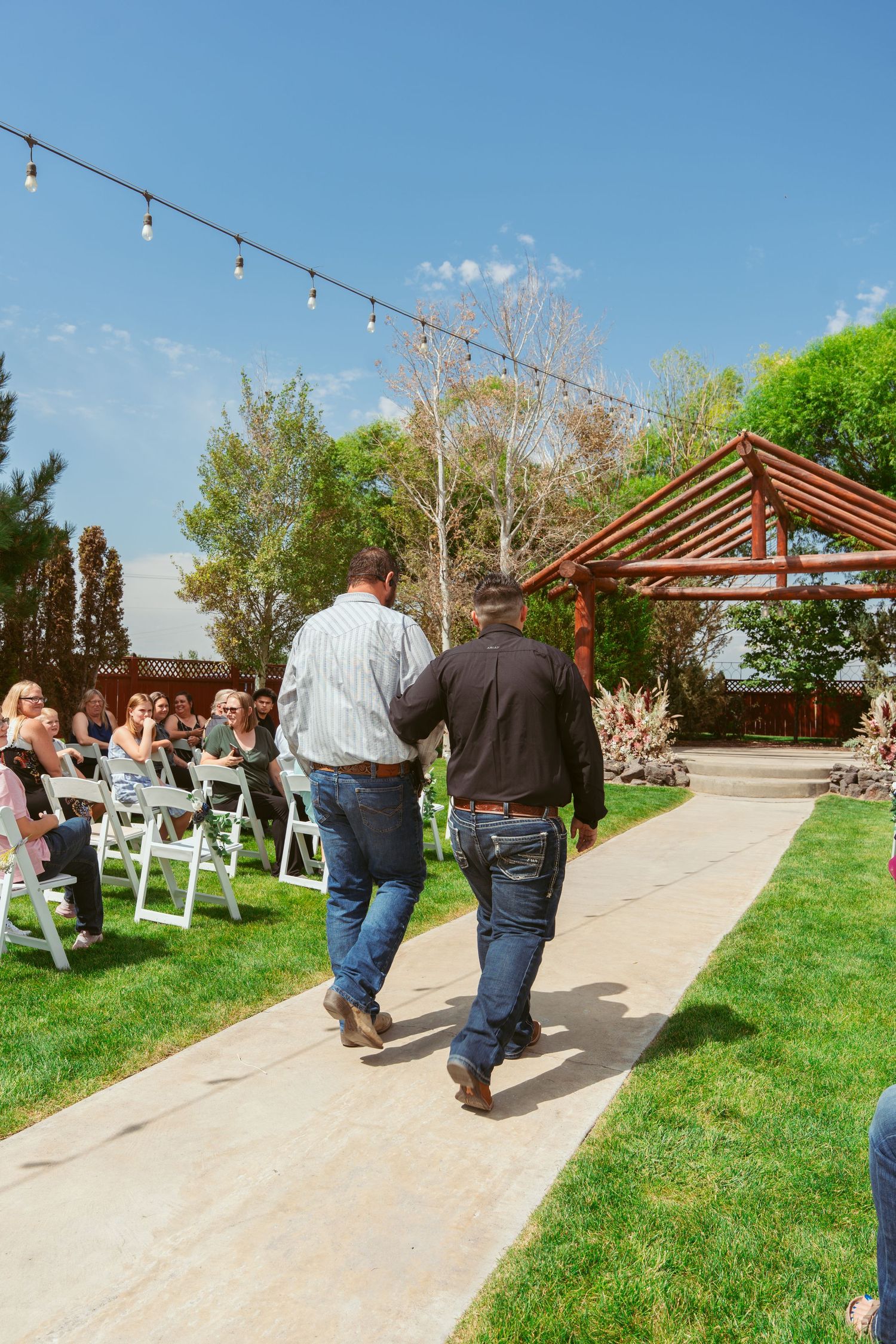 Outdoor venue with wooden pergola, string lights, and concrete walkway leading to ceremony area.